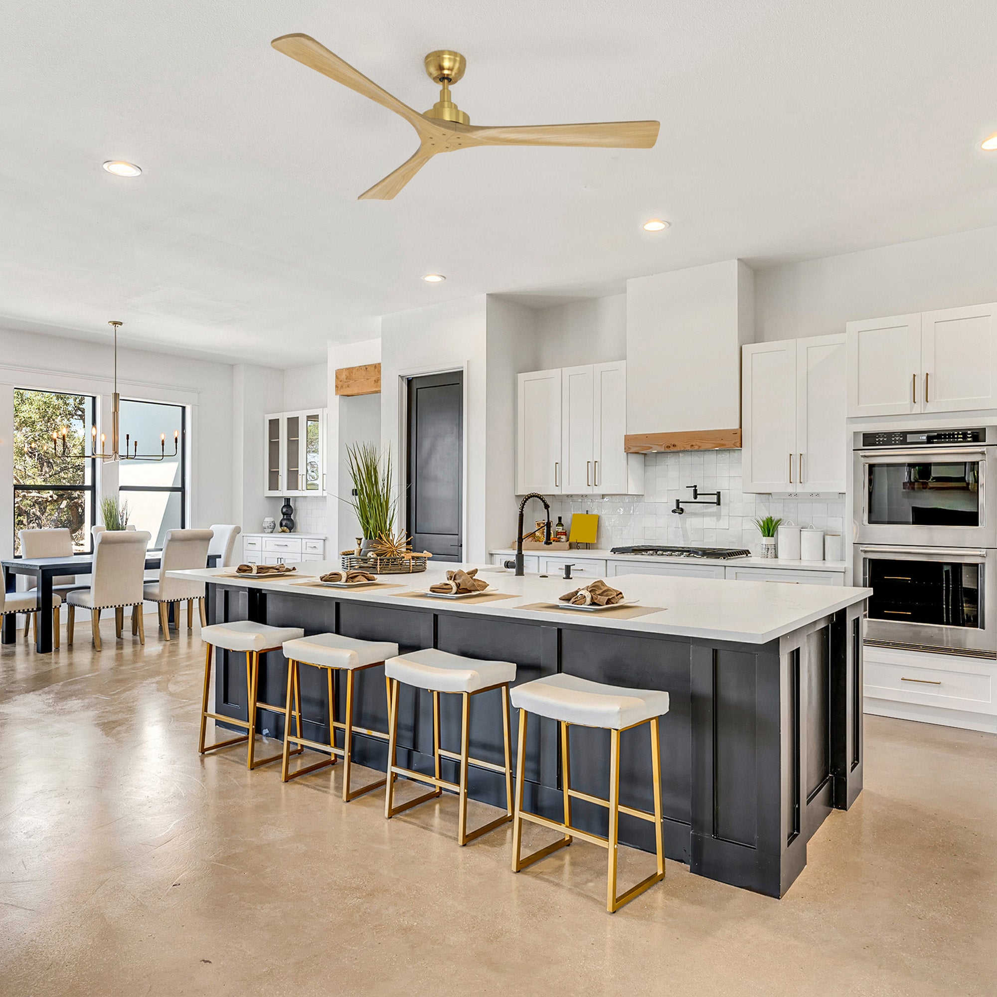 Smafan Thora 60 inch wood ceiling fan with remote in a gold finish, mounted above a large gray kitchen island with white countertop and gold bar stools. The modern open-concept kitchen features white cabinets, stainless steel appliances, a wood range hood, and a dining table in the background.