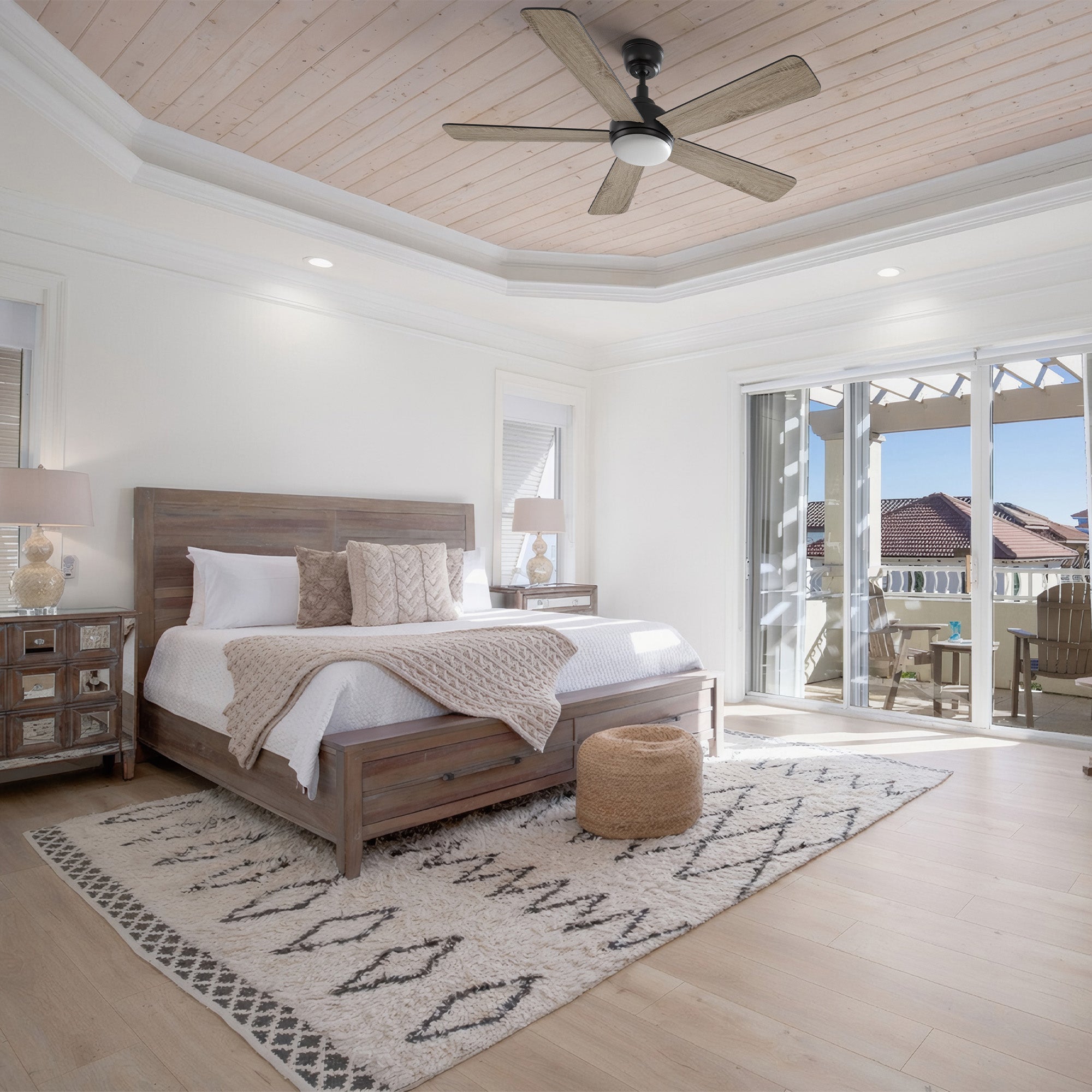A cozy rustic bedroom featuring the Smafan Toplam 52-inch wood ceiling fan with light mounted on a light wood plank ceiling. The room includes a large bed with a wood frame and white comforter, patterned area rug, woven ottoman, and matching nightstands with lamps. Large glass doors open to a bright balcony with outdoor seating.