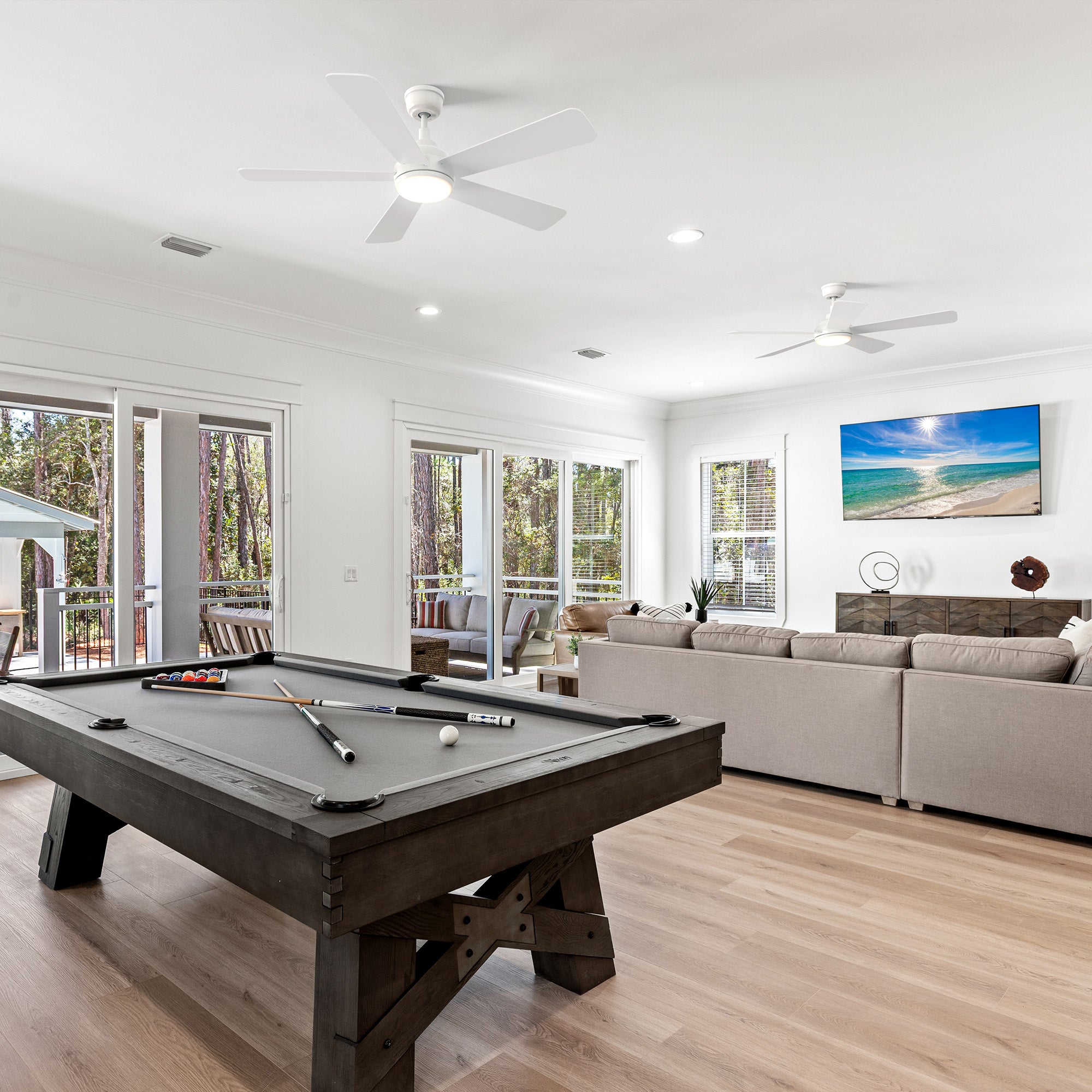 A modern open-concept living space featuring two Smafan Toplam 52-inch white ceiling fans with lights on the ceiling. The room includes a dark wood pool table with a gray felt top, a large beige sectional sofa facing a wall-mounted TV, and expansive glass doors revealing a forest-view deck with patio furniture.