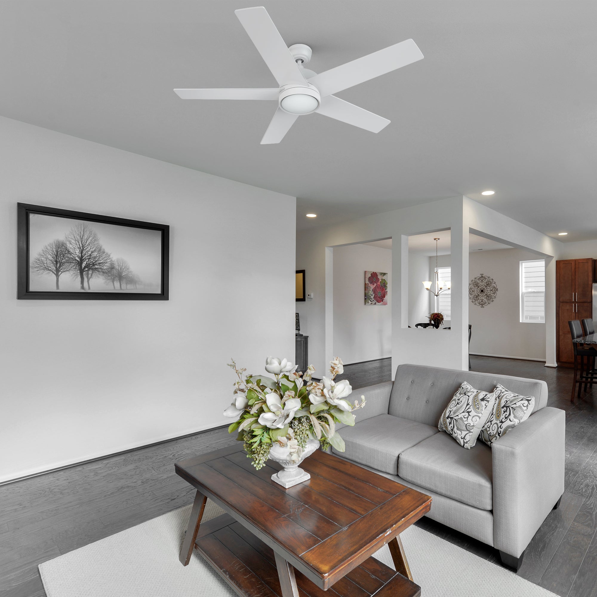 A minimalist living room featuring a 52-inch Smafan Trafford2 white ceiling fan with light, mounted on the ceiling. The room includes a grey sofa with patterned pillows, a wooden coffee table with a vase of white flowers, and a black-and-white tree photograph on the wall. The space has dark hardwood floors, a white rug, and an open layout with large windows, a chandelier, and modern decor in the background.