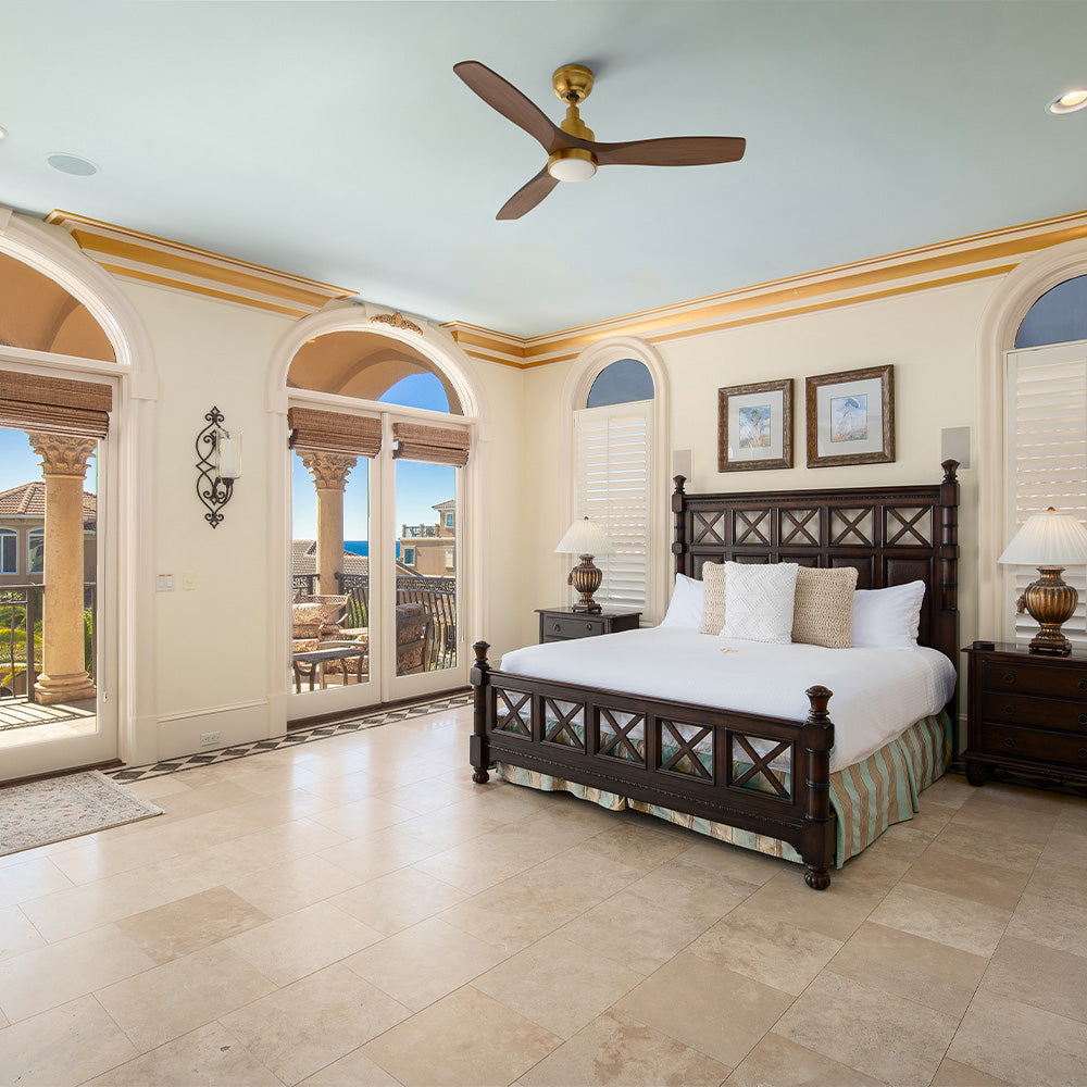 A luxurious bedroom with a large dark wood four-poster bed and marble floors, featuring arched glass doors leading to a balcony with an ocean view, and a Smafan Ultiem 48-inch wood ceiling fan in a gold finish with LED light mounted on the light blue ceiling.