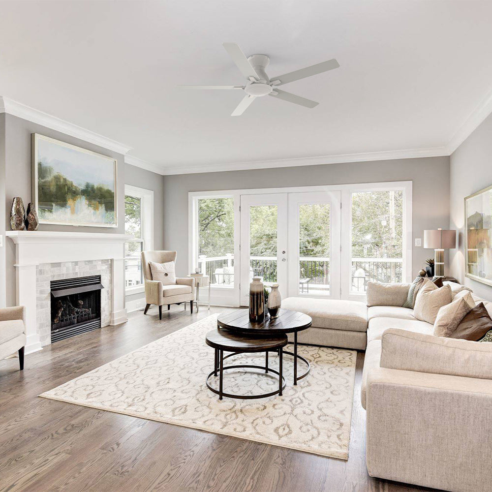 A bright, modern living room featuring light grey walls and hardwood floors, centered around a white fireplace with a marble-tile surround and a painting mounted above. The room is furnished with a large beige sectional sofa, a wingback armchair, and two round, nesting coffee tables with dark wood tops and black metal bases. A large area rug sits on the floor, and a white ceiling fan with five blades is visible overhead. Large glass doors lead out to a deck or patio.