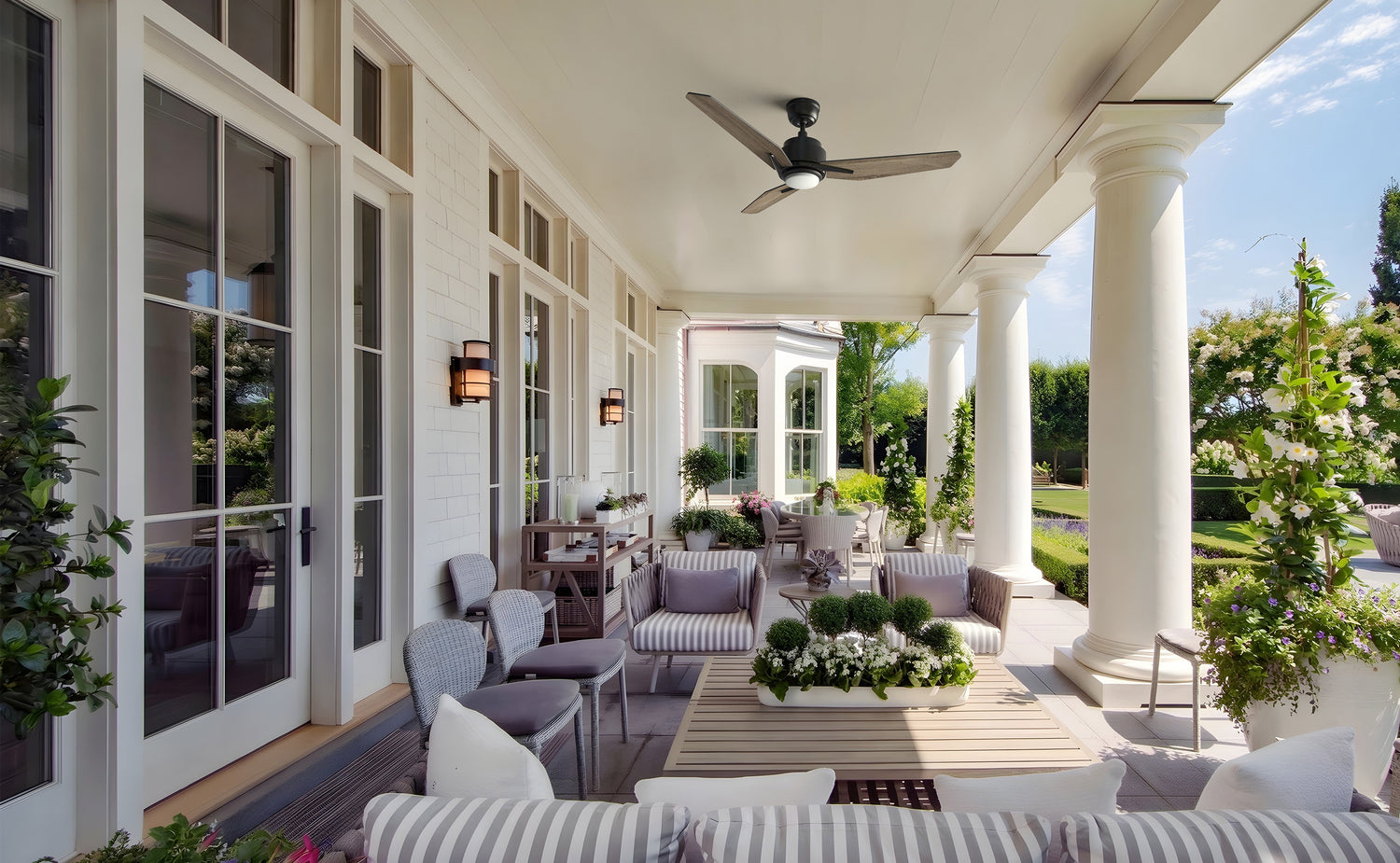 The scene includes modern grey striped lounge chairs and a wooden coffee table centered around a lush white floral arrangement. A black ceiling fan hangs from the porch roof, while large glass doors and manicured gardens frame the background.