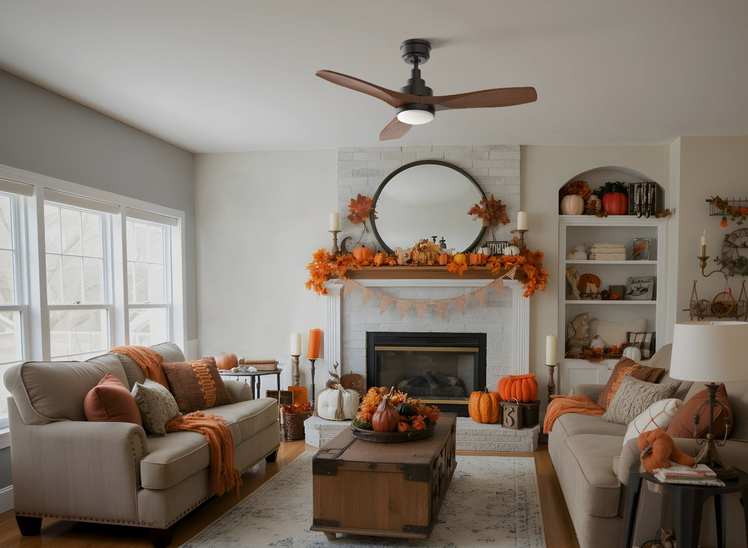 A cozy autumn-themed living room decorated for Halloween, featuring the Smafan Ultiem 48-inch ceiling fan with warm wood-toned blades overhead. The space includes a white brick fireplace with a round black-framed mirror, a garland of orange leaves, pumpkins, and candles on the mantel. Two light-colored sofas with orange pillows face each other around a wooden trunk coffee table topped with pumpkins.