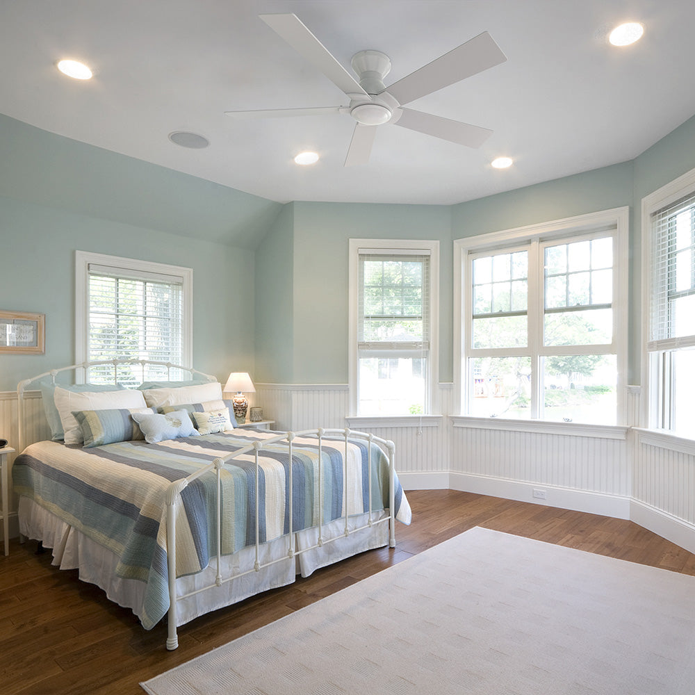 A bedroom with light blue walls and white wainscoting. A white metal bed frame with a blue and white striped comforter is in the foreground, and a large bay window with white blinds overlooks a green outdoor area. A Vetric 52 inch Low Profile Ceiling Fan in white without light is mounted on the ceiling, surrounded by recessed lighting.