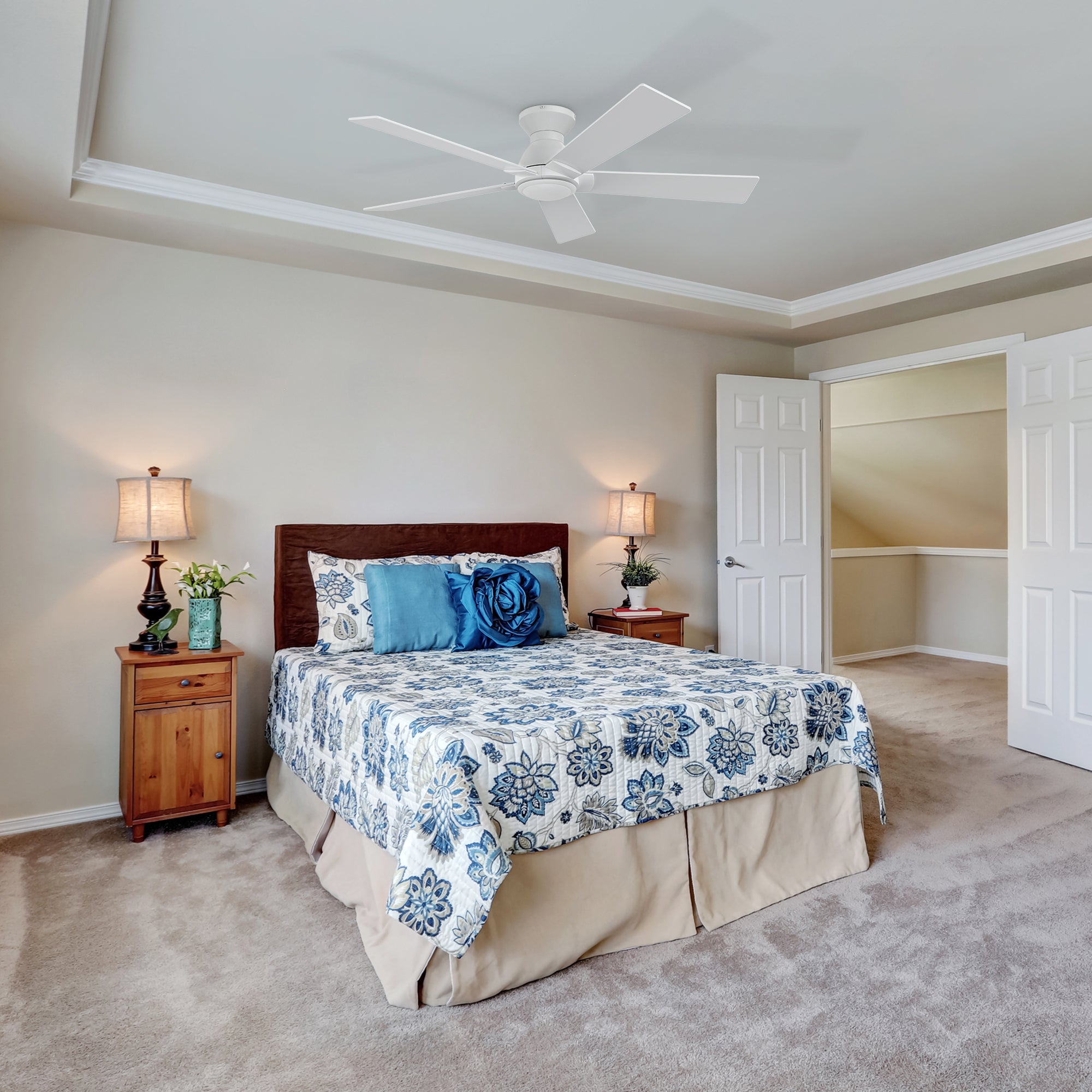 A bedroom featuring a 52-inch low-profile white ceiling fan mounted on the ceiling, with a bed adorned with a blue floral comforter, blue pillows, and a beige bed skirt. The room includes two wooden nightstands, each with a lamp and decorative items, light beige walls with a tray ceiling, carpeted flooring, and an open door leading to another room.