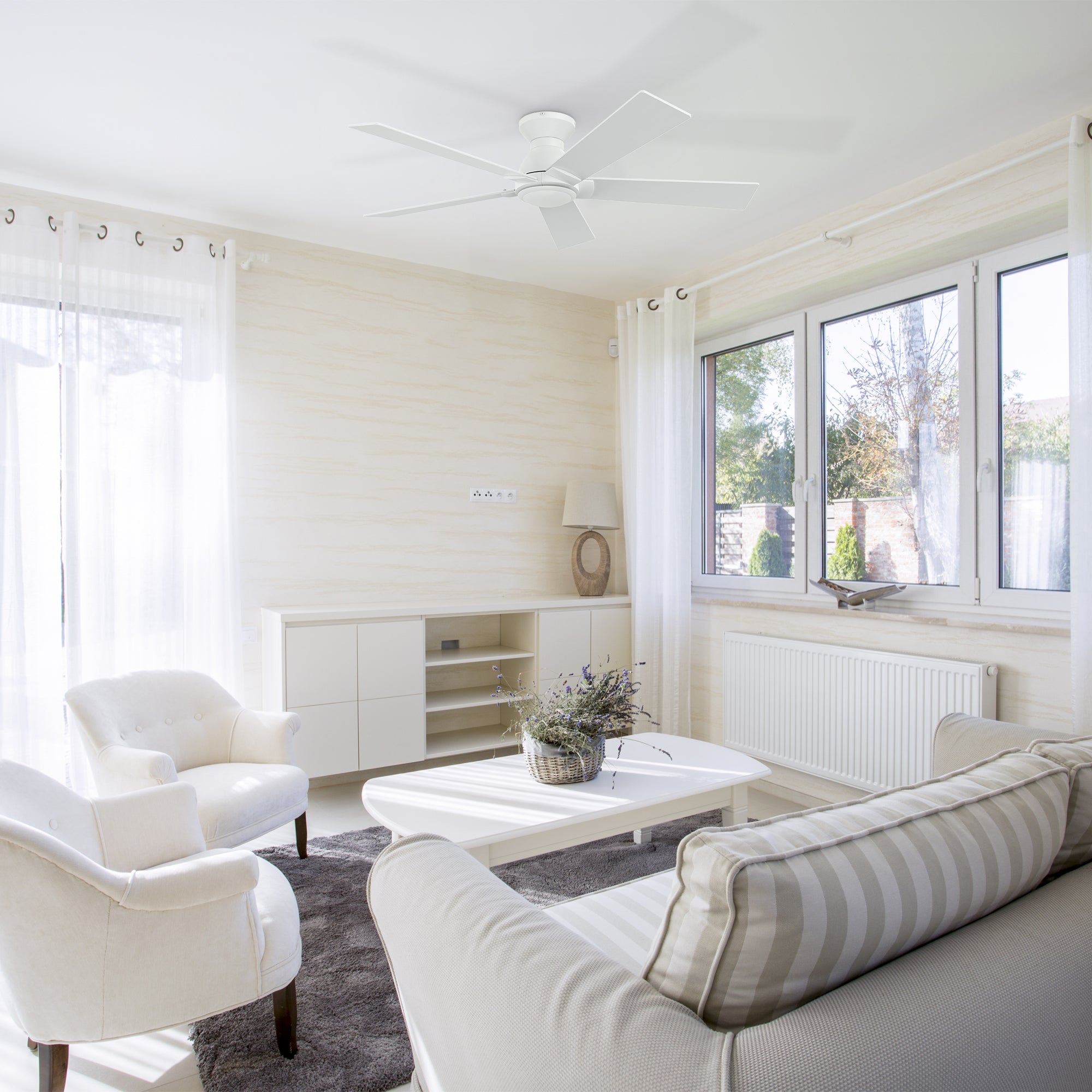 A bright living room featuring a 52-inch low-profile white ceiling fan mounted on the ceiling, with two white armchairs, a white sofa with striped cushions, and a white coffee table. The room includes a white cabinet with decorative items, sheer curtains, large windows with natural light, a radiator, and a gray area rug on the light wood floor.