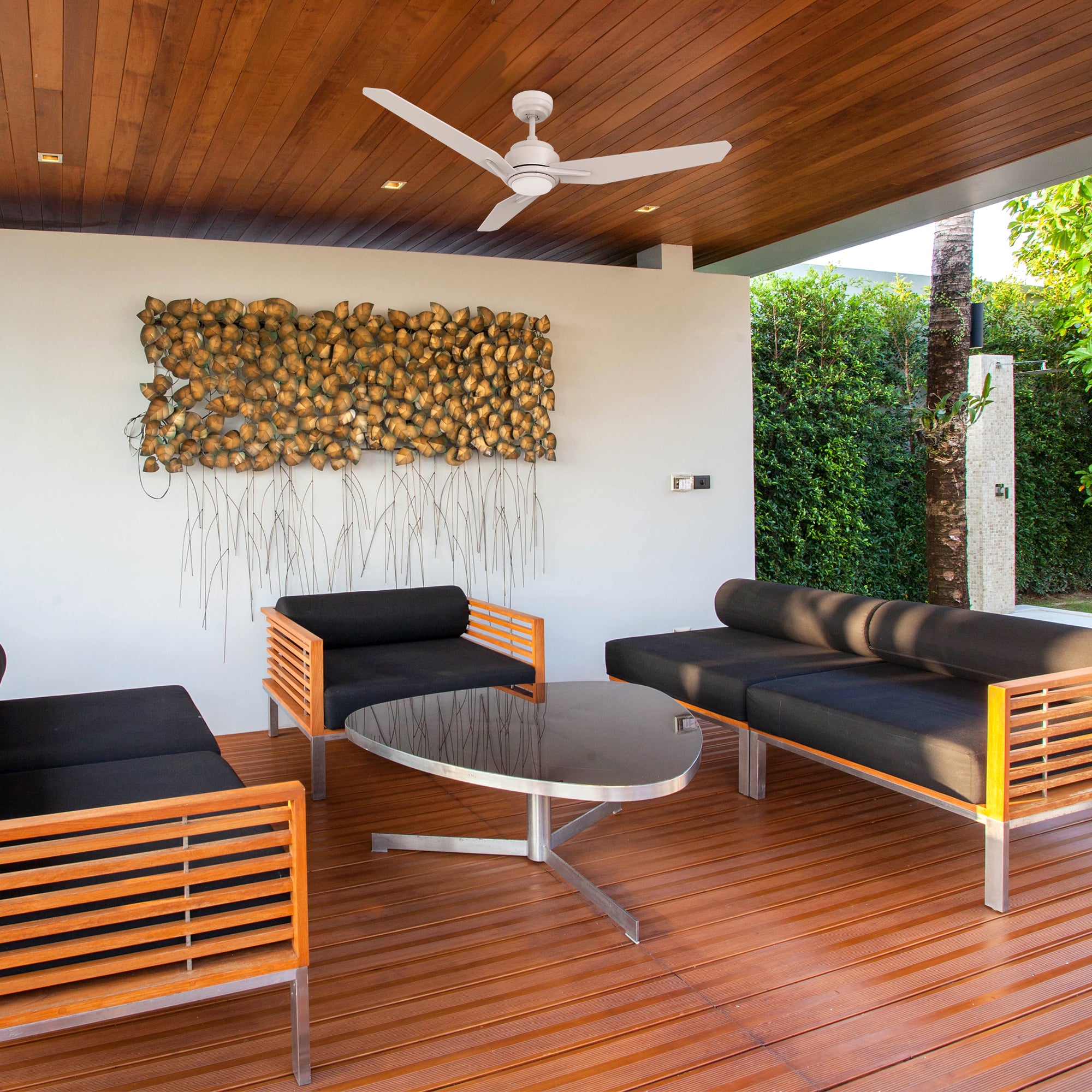 Modern covered patio with Smafan white 56-inch Tilbury outdoor ceiling fan featuring 3 blades. The space includes black cushioned wooden seating, a sleek round coffee table, and a sculptural wall art installation.