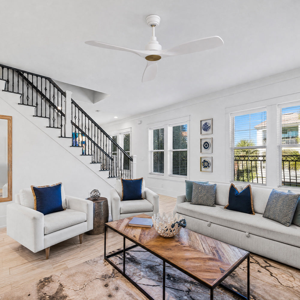 An airy living room featuring a smart white ceiling fan without a light, accented with a gold motor hub. The space includes a white sofa with navy blue pillows, a wood chevron-patterned coffee table, and a black wrought-iron staircase against a white wall.