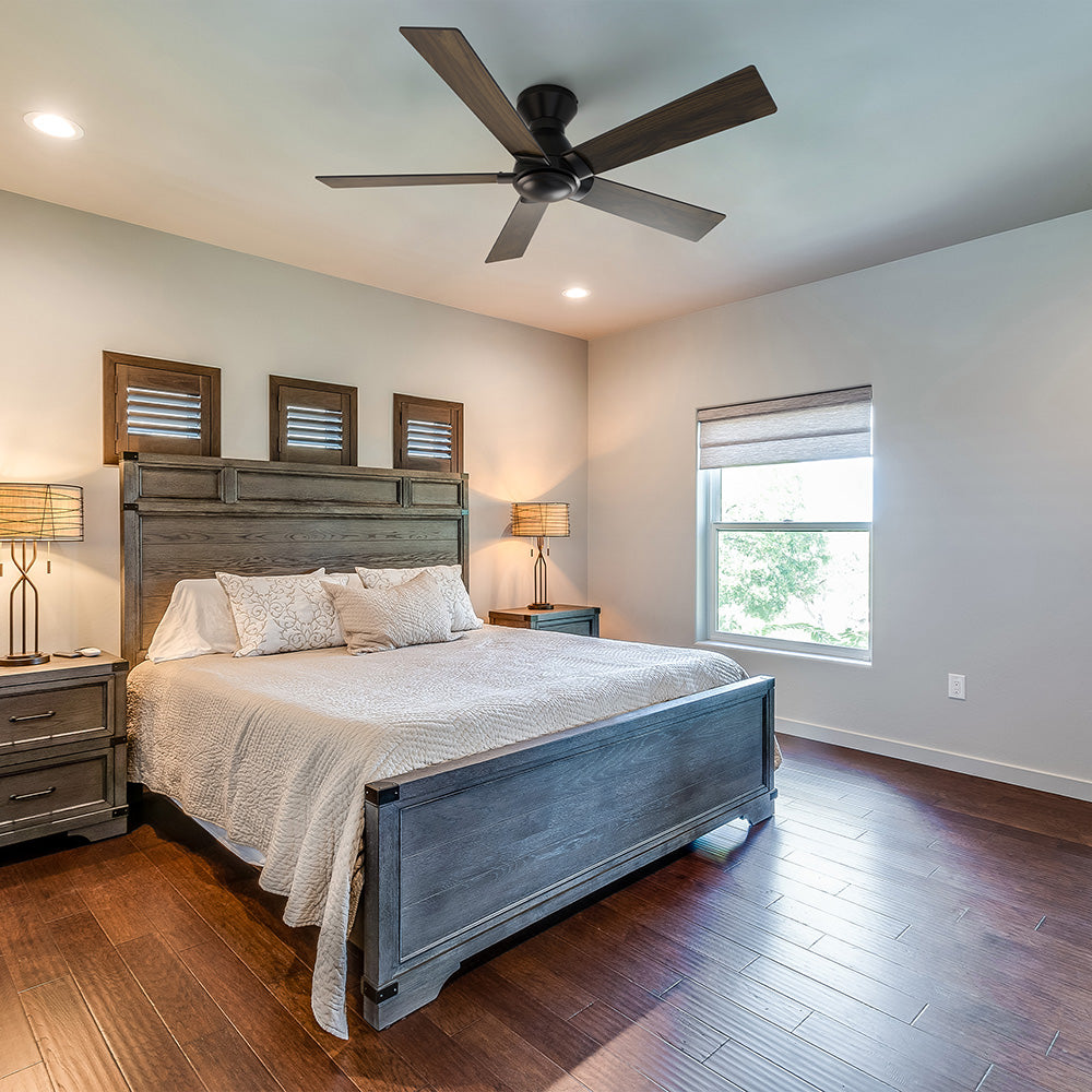 A modern bedroom featuring a gray wooden bed frame with a matching headboard and two nightstands with lamps. A large window with a shade is on the right, and above the bed are three small windows with wooden shutters. A Vetric 52 inch Low Profile Ceiling Fan without light hangs from the ceiling, and the room has dark wood flooring.