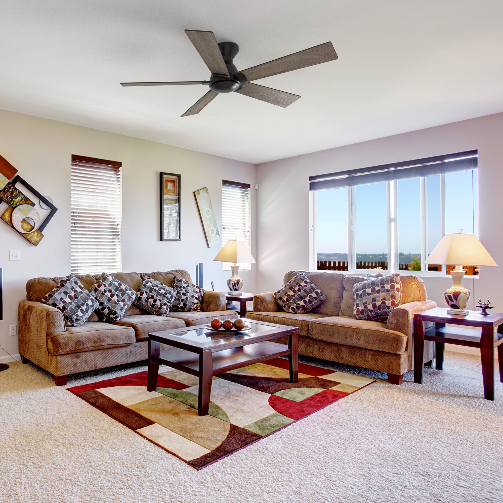 A living room features a Vetric 52 inch Low Profile Ceiling Fan without light mounted on the ceiling, positioned above a brown sectional sofa and a dark wood coffee table. The room is carpeted in a light color and includes a colorful geometric rug under the coffee table. Large windows with blinds offer a view of an outdoor landscape, and the room is decorated with wall art and table lamps on end tables.