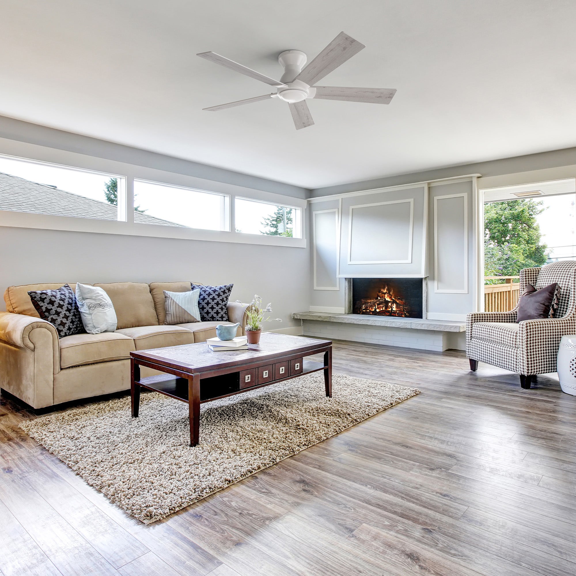 A modern living room featuring a Vetric 52 inch Low Profile Ceiling Fan with Remote No Light, positioned centrally on the white ceiling. The room includes a beige sofa with patterned pillows, a dark wood coffee table on a shag rug, a fireplace with a light gray mantel, and large windows offering a view of trees.