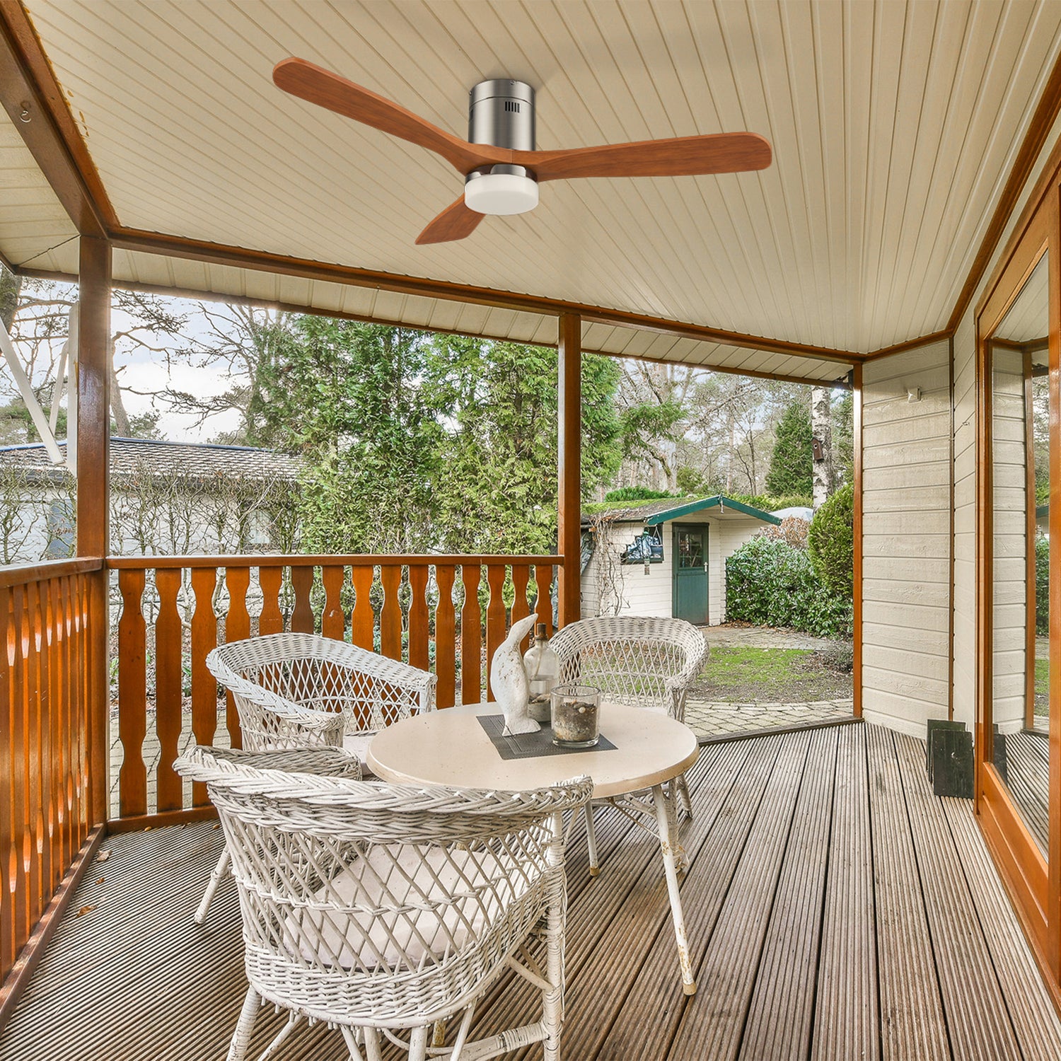 Covered patio with white wicker chairs and a round table set with decor items. An 52 inch outdoor flush mount ceiling fan with light and wooden blades hangs above. Garden and shed are visible beyond the patio railing.