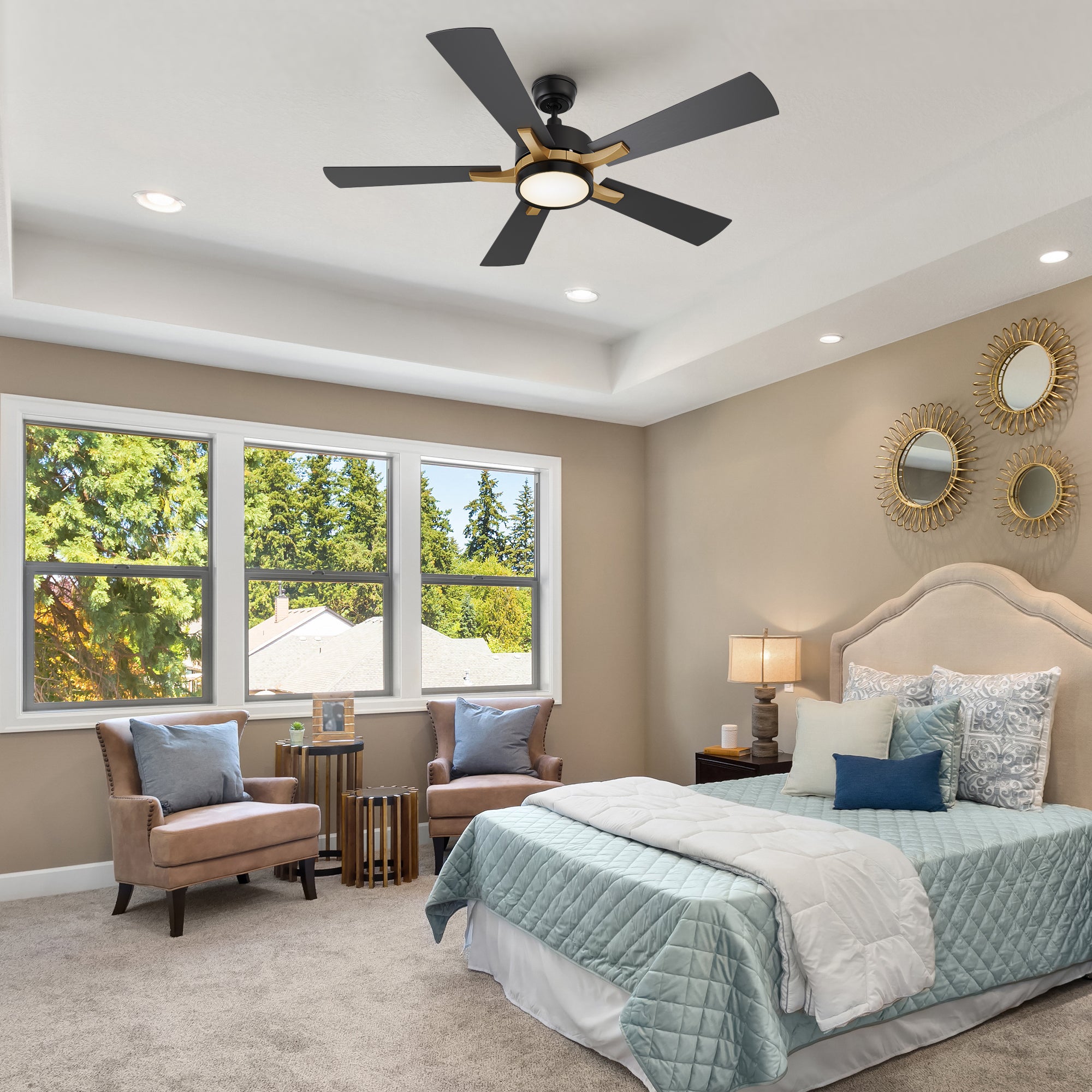 A bright and cozy bedroom featuring neutral-toned walls, a tufted beige headboard, and coordinated bedding in soft blue and white. The room includes accent chairs by the window, nightstands with lamps, and decorative wall mirrors. A Smafan Apex 52-inch smart ceiling fan with LED light is mounted on the tray ceiling, adding comfort and a modern touch.