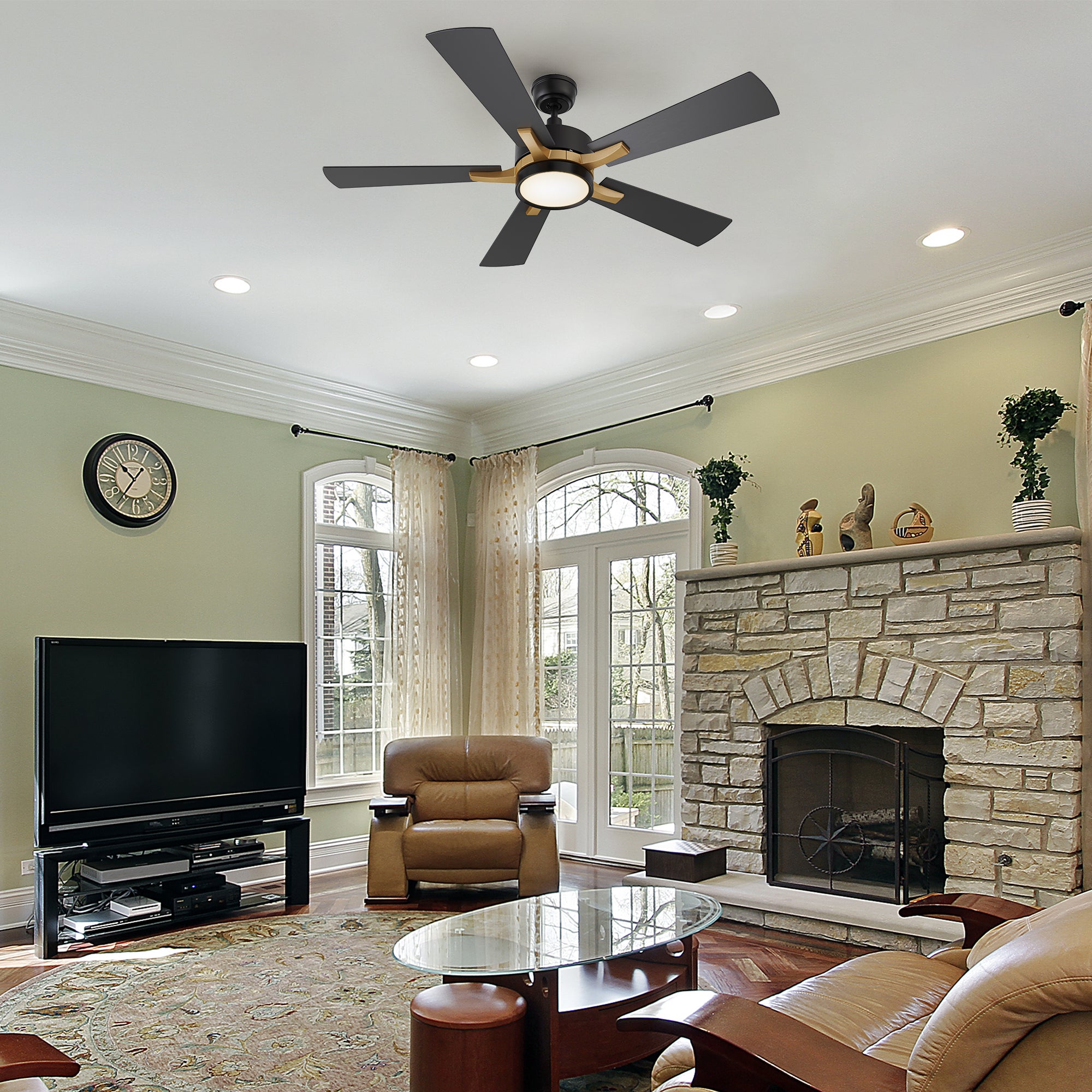 A cozy, well-lit living room with sage green walls, a stone fireplace, and modern furnishings including a leather armchair, glass coffee table, and flat-screen TV. The 52-inch Smafan Apex smart ceiling fan with LED light is mounted at the center of the ceiling, offering smart control and sleek design that complements the space.