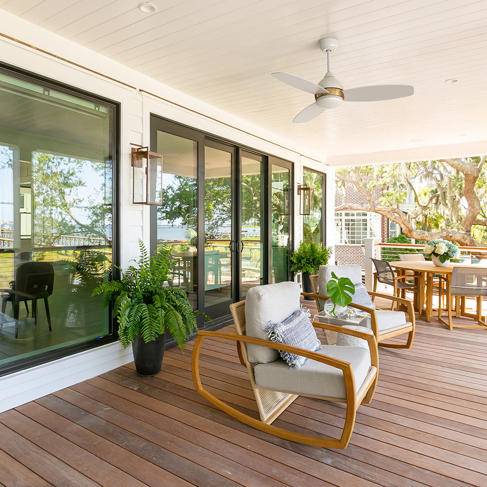 Control this smart home enabled outdoor Carro Bretton 48 inch modern white outdoor ceiling fan with downrod, gold accent, and light kit installed on a spacious covered patio deck with rocking chairs and dining area.