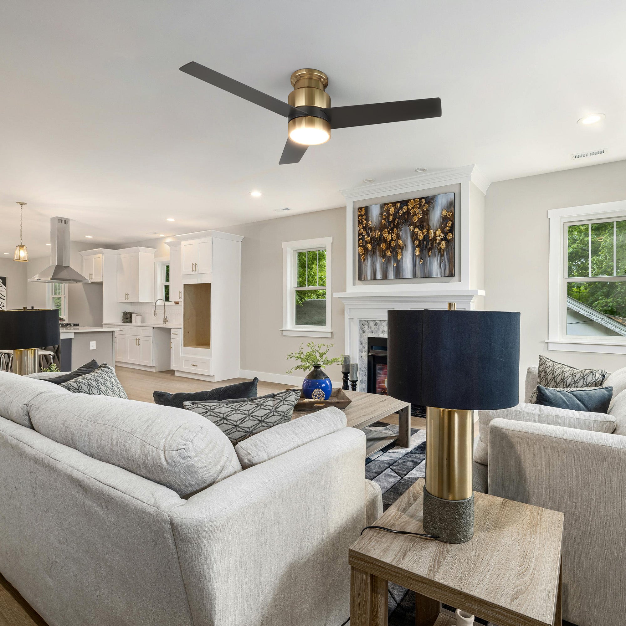 An open-concept living room and kitchen featuring a light grey sectional sofa with throw pillows, a wooden coffee table, and a matching side table with a gold-based lamp and dark blue shade. The room includes a fireplace with abstract artwork above it, and is illuminated by a flush mount ceiling fan in black and gold with an energy-efficient LED light kit, all styled in a modern farmhouse aesthetic with a neutral color palette.