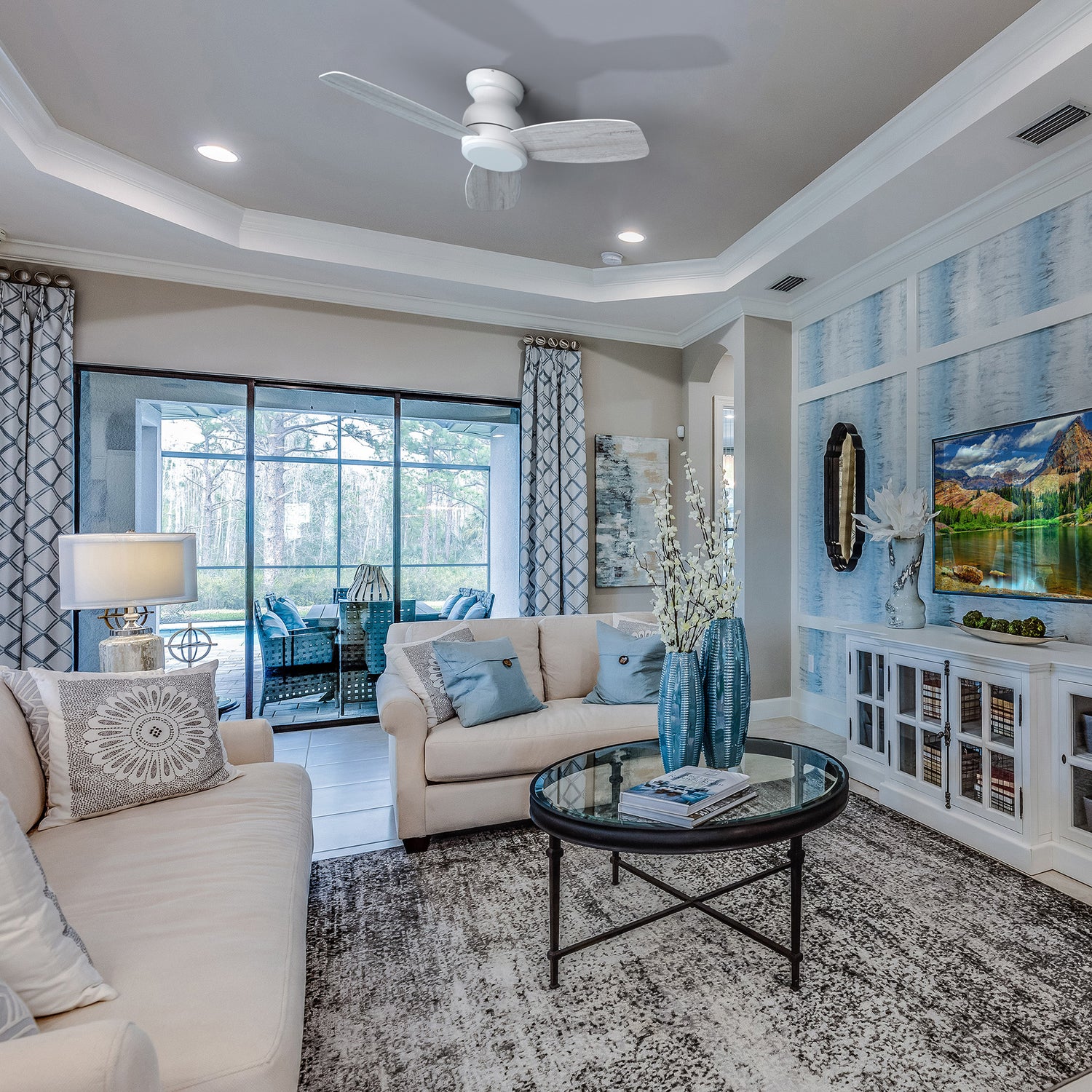 A modern living room featuring a white Osborn 44-inch flush mount ceiling fan without a light, above a glass coffee table and a patterned rug. The room includes a light-colored sofa with throw pillows, a large window looking out onto a patio, and a wall with blue and white patterned wallpaper.