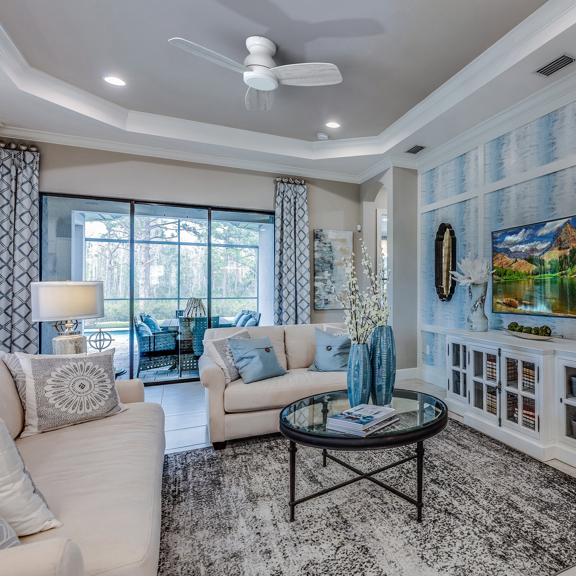 A modern living room featuring a white Osborn 44-inch flush mount ceiling fan without a light, above a glass coffee table and a patterned rug. The room includes a light-colored sofa with throw pillows, a large window looking out onto a patio, and a wall with blue and white patterned wallpaper.