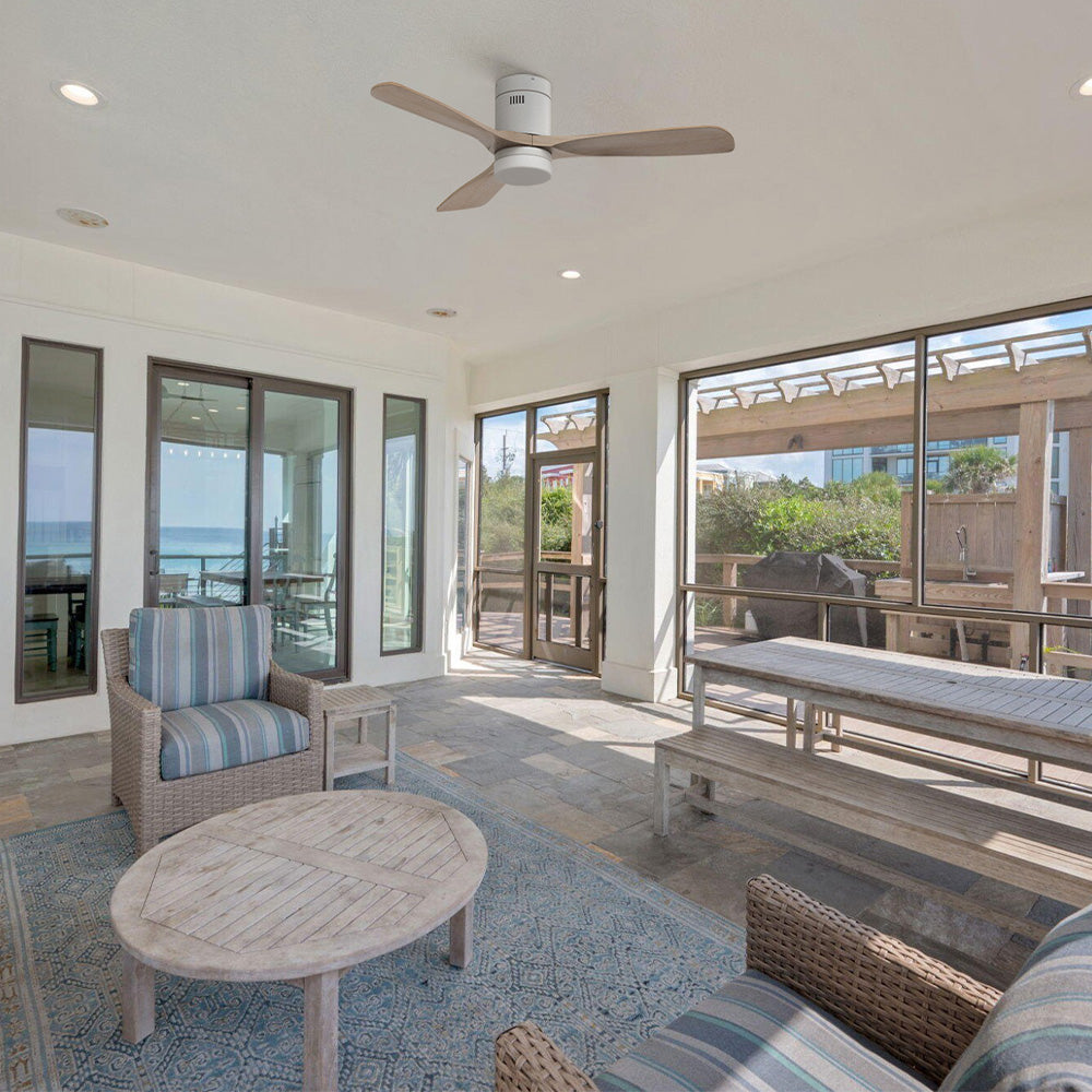 Smafan Alfa 52 inch low profile ceiling fan with 3 light wood blades installed in a bright coastal-style sunroom. The modern indoor-outdoor ceiling fan complements the airy seating area, large windows, and beach-inspired décor while providing comfortable airflow and a clean, minimalist look.