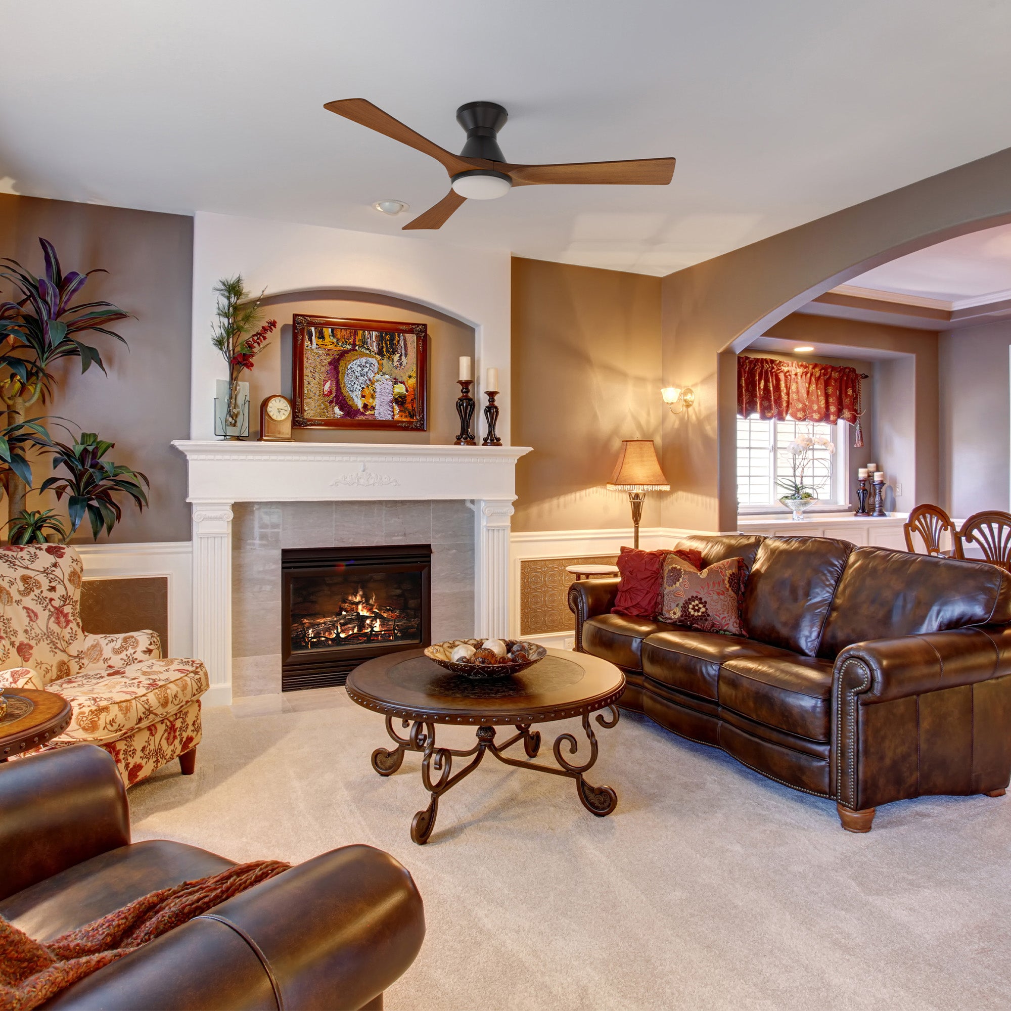 Cozy living room with brown leather sofas, round coffee table, floral armchair, fireplace with art above, plants, and warm earth-toned walls. A low profile smart ceiling fan with light and solid wood 3 blades accents the carpeted floor and living room chill zone view.