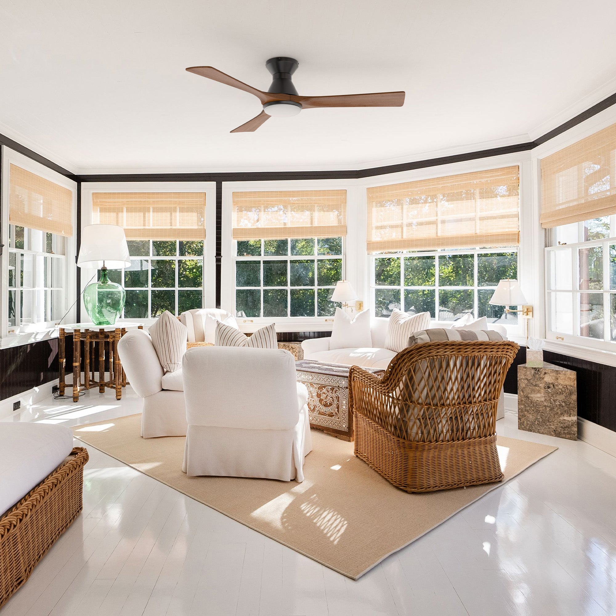 Bright sunroom with large windows, white walls, wicker and white upholstered furniture, a beige rug, and a low-profile smart ceiling fan with light; sunlight streams onto glossy white floors.