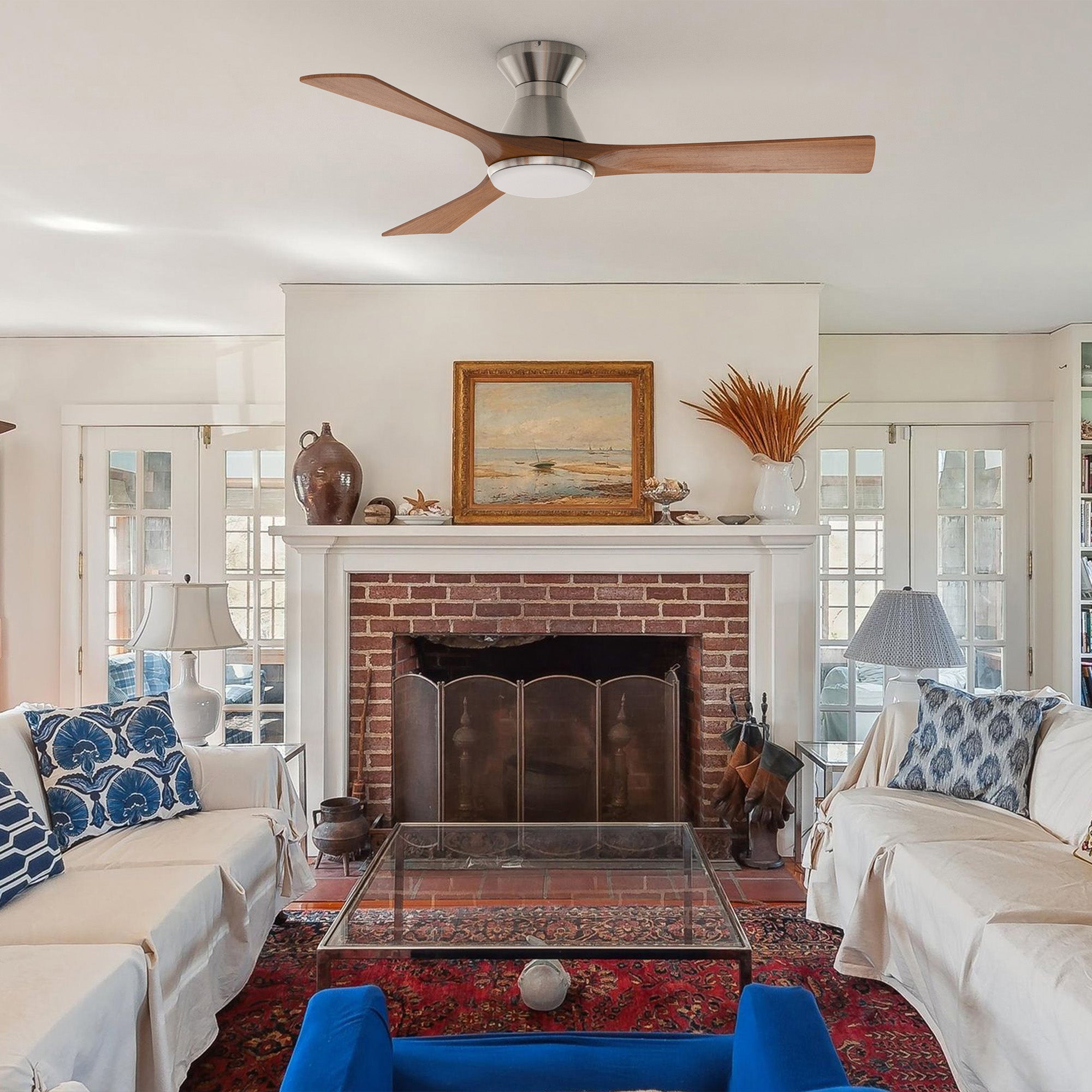 A cozy rustic living room with a brick fireplace, glass coffee table, white sofas with blue patterned pillows, and a blue chair. A low profile ceiling fan with light and wooden blades is centered above, while art and vases decorate the mantel.