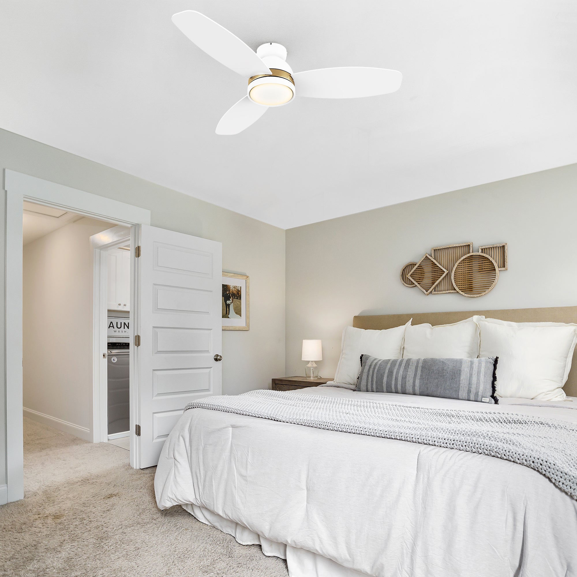 Modern bedroom with 52 inch Biscay flush mount ceiling fan in white and gold featuring a gold accent ring and integrated LED light, mounted above a neatly made bed with white bedding, gray throw pillow, and neutral wall decor.