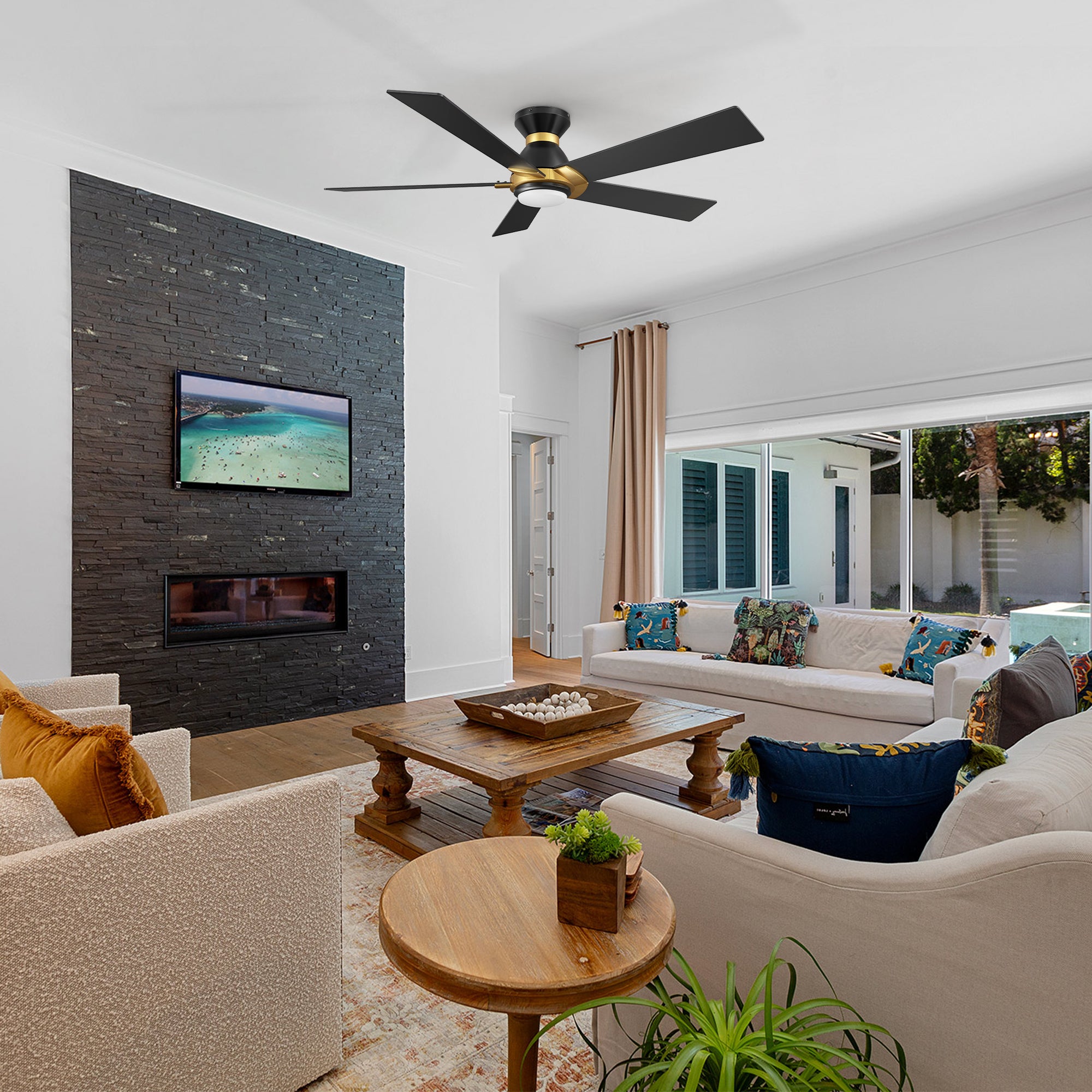 A modern living room featuring a smart low-profile Aspen 52-inch ceiling fan with dimmable LED light, mounted on the ceiling. The room includes a stone-accented wall with a mounted TV and fireplace, a beige sofa with colorful pillows, a wooden coffee table with decorative items, a small round side table with a plant, and large windows opening to an outdoor pool area.