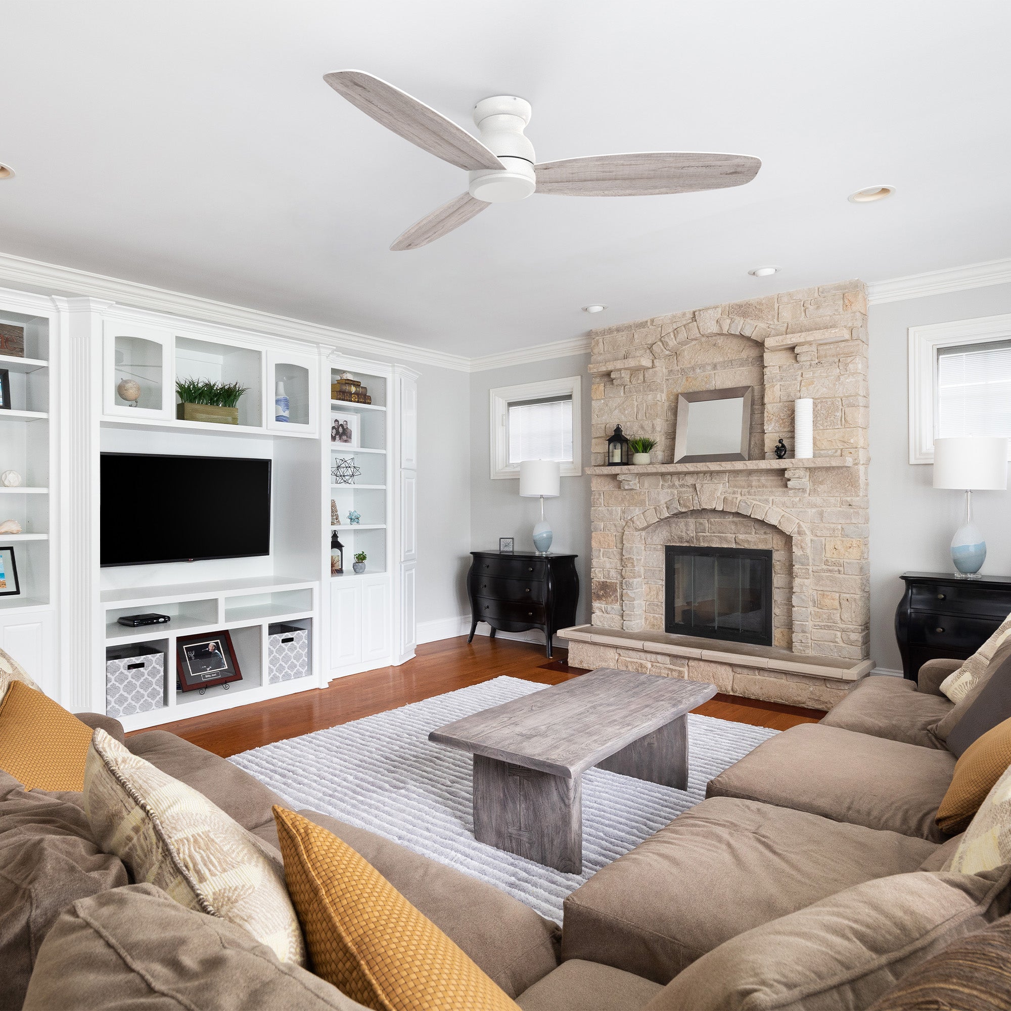 A bright living room with a large sectional sofa, wooden coffee table, stone fireplace, built-in shelves with decor, TV, and a low profile ceiling fan in white. The room features neutral colors and natural light from small windows.