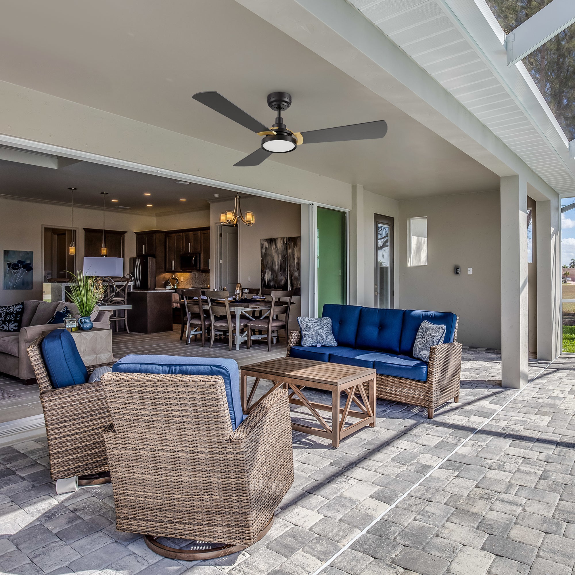 Modern covered patio with rattan furniture and blue cushions, featuring a black 52-inch Smafan Carro Viter smart outdoor ceiling fan with integrated LED light, installed overhead for comfortable airflow. The space opens into a stylish kitchen and dining area.