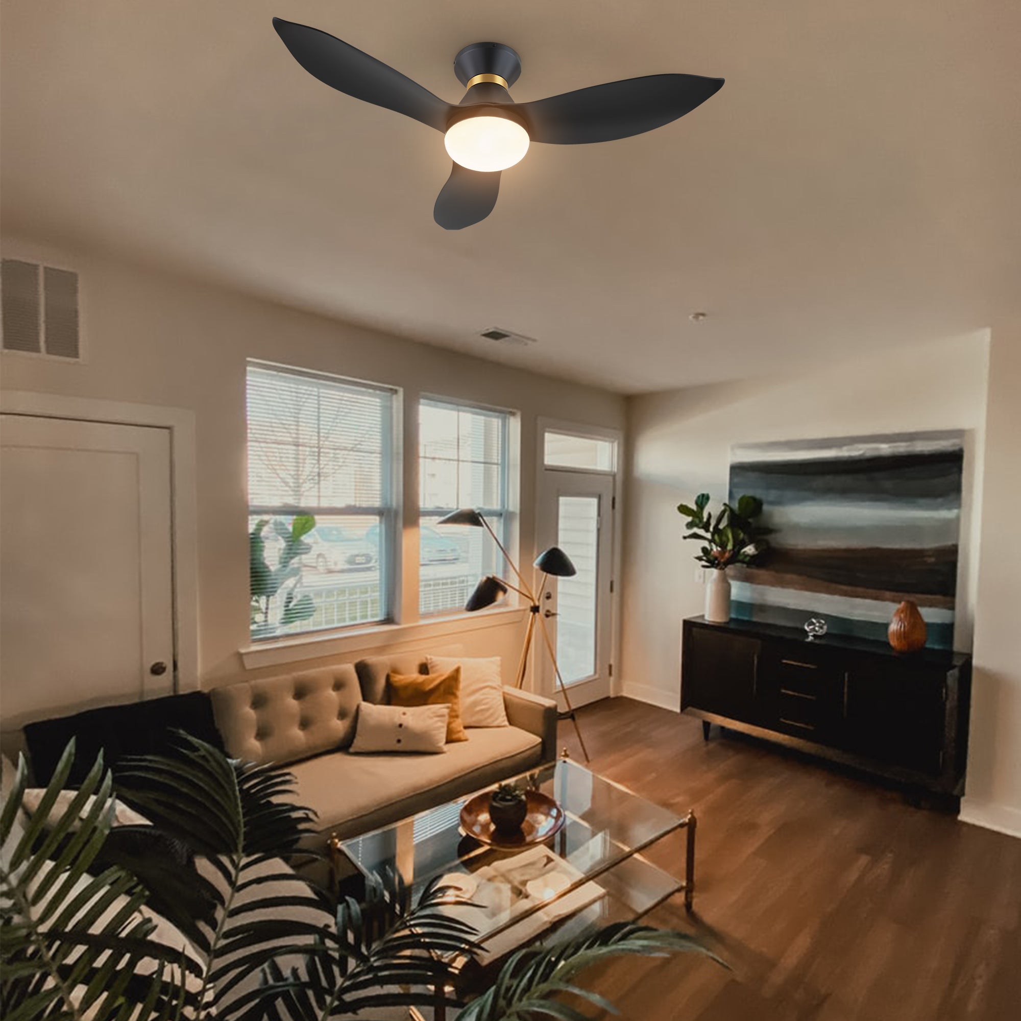A modern living room featuring a black and gold flush-mount smart ceiling fan with three blades and an integrated light, mounted on a beige ceiling. The room includes a beige sofa with cushions, a glass coffee table with plants, a dark wood cabinet with decor, large windows with blinds, and a hardwood floor.