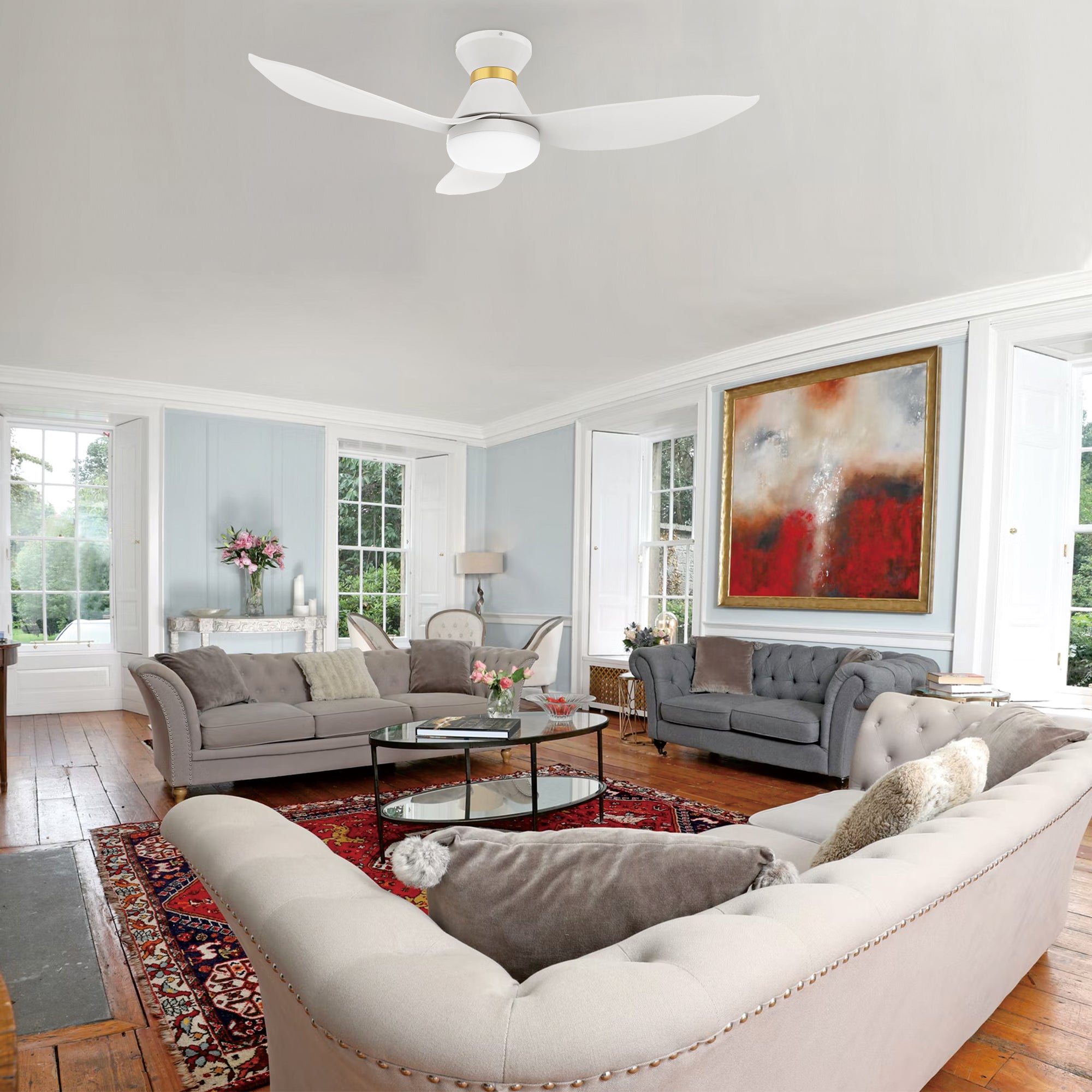 A stylish living room with a 45-inch Joliet flush-mount smart ceiling fan in white and gold with an integrated light, mounted on a white ceiling. The room features two tufted sofas with gray and beige cushions, a glass coffee table, a red patterned rug, a large red and gold framed painting, windows with natural light, and wooden flooring.