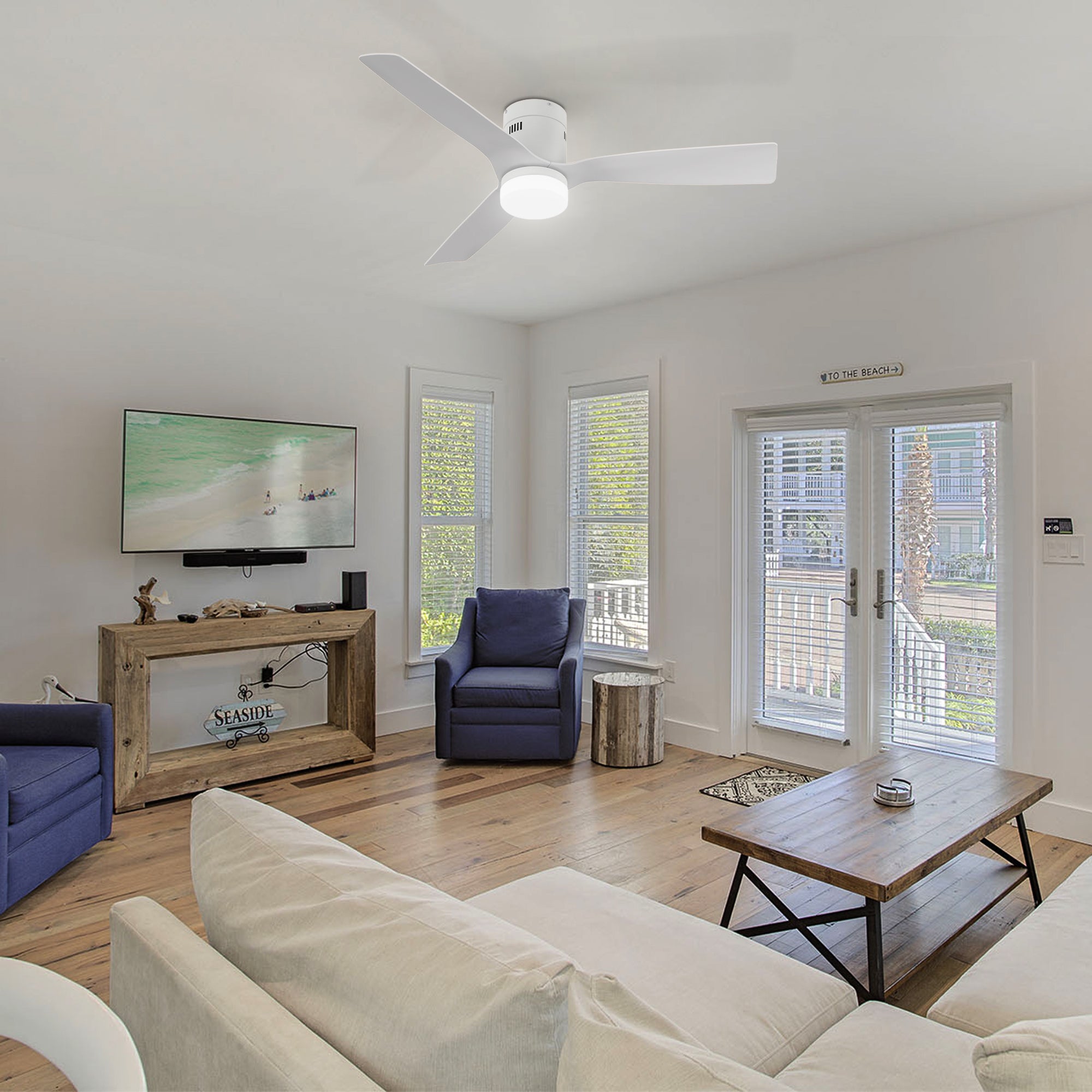 A modern living room featuring a white ceiling fan with light mounted on the ceiling, surrounded by two blue armchairs, a white sectional sofa, and a wooden coffee table with a metal base. The room includes a wooden TV stand with a flat-screen TV displaying a beach scene, white wooden blinds on large windows, a glass door leading to a balcony, and hardwood flooring with a small area rug.