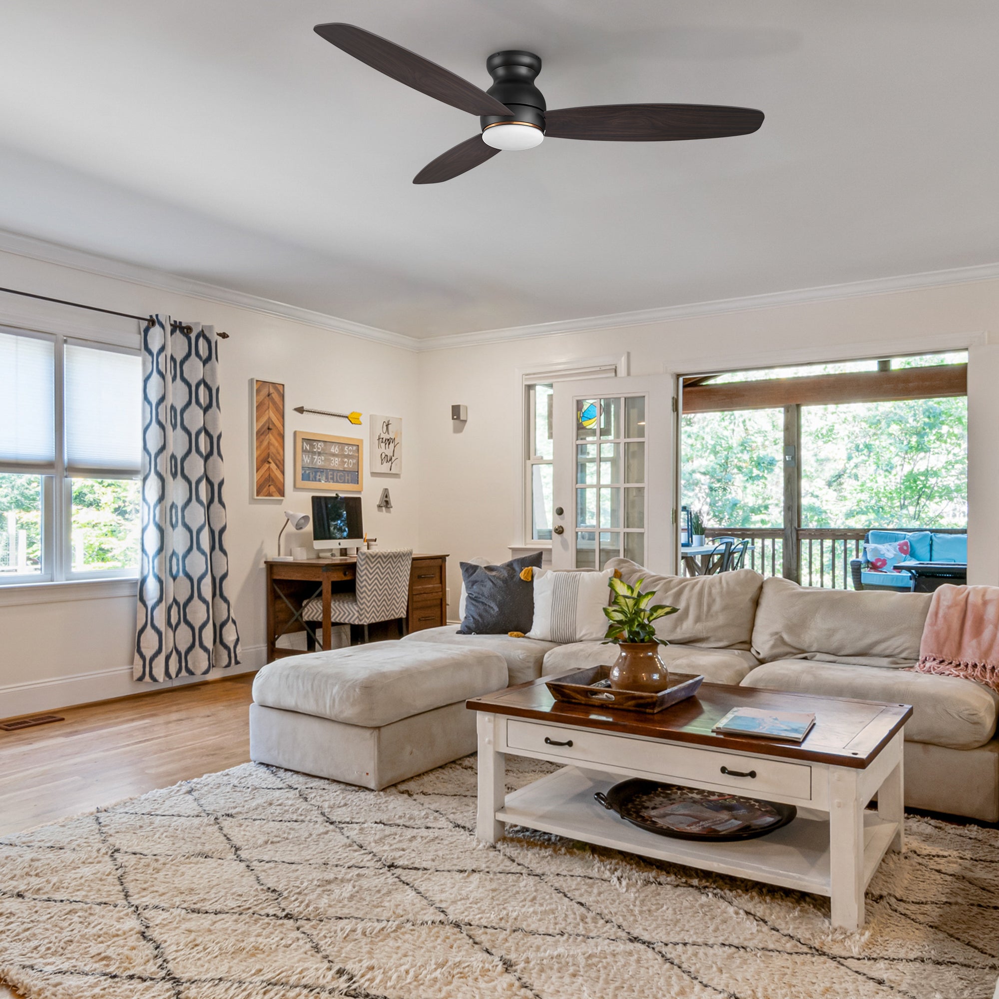 A cozy living room with a sectional sofa, patterned rug, coffee table, desk, and 60 inch low profile smart ceiling fan with light. Large windows and a door open to a sunroom with a view of greenery outside.