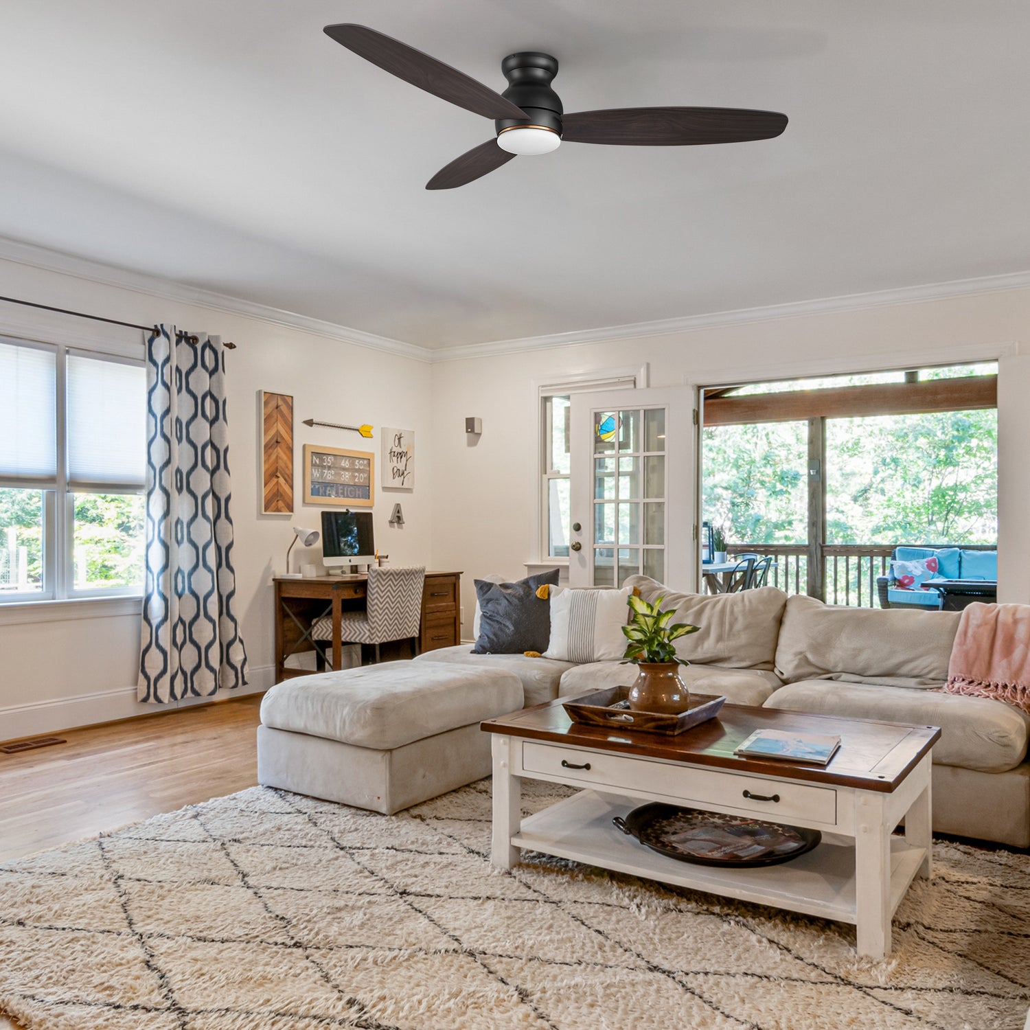 A cozy living room with a sectional sofa, patterned rug, coffee table, desk, and 60 inch low profile smart ceiling fan with light. Large windows and a door open to a sunroom with a view of greenery outside.