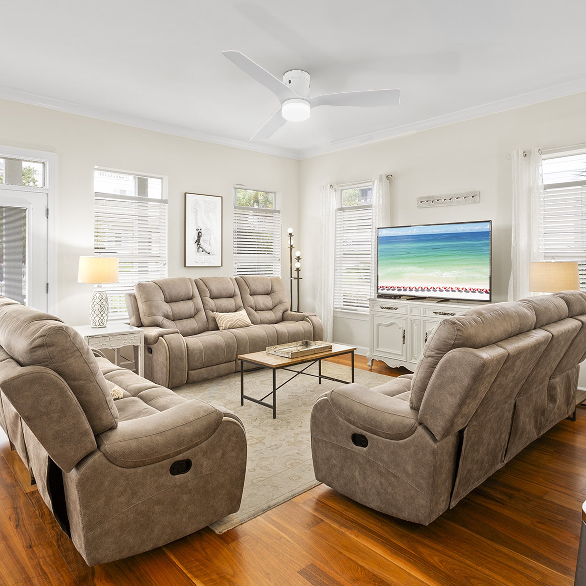 A modern living room featuring a 52-inch Smafan Striver flush-mount smart ceiling fan with light in white, mounted on the ceiling. The room includes two tan reclining sofas with cushions, a black metal and wood coffee table, a white cabinet with a TV displaying a beach scene, white curtains, wooden blinds, two table lamps, a side table, and hardwood flooring with a light-colored area rug.