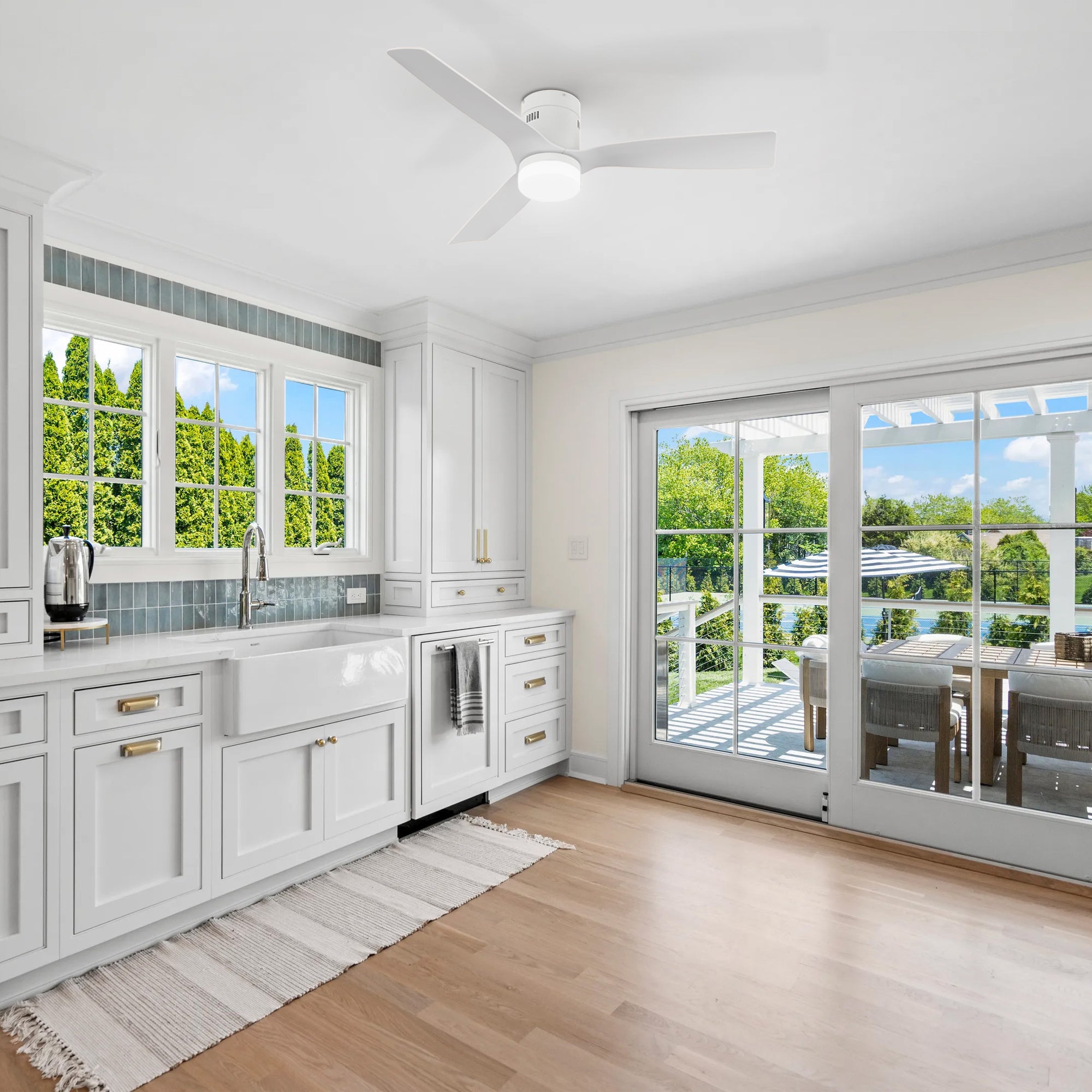 A rustic kitchen featuring a 52-inch Smafan Striver white low-profile ceiling fan with light, mounted on the ceiling. The kitchen includes white cabinetry with gold handles, a white farmhouse sink with a blue tile backsplash, a coffee maker on the counter, and a striped rug on hardwood flooring. Large windows and a glass sliding door offer views of a lush green outdoor area with a dining table and chairs under a pergola.