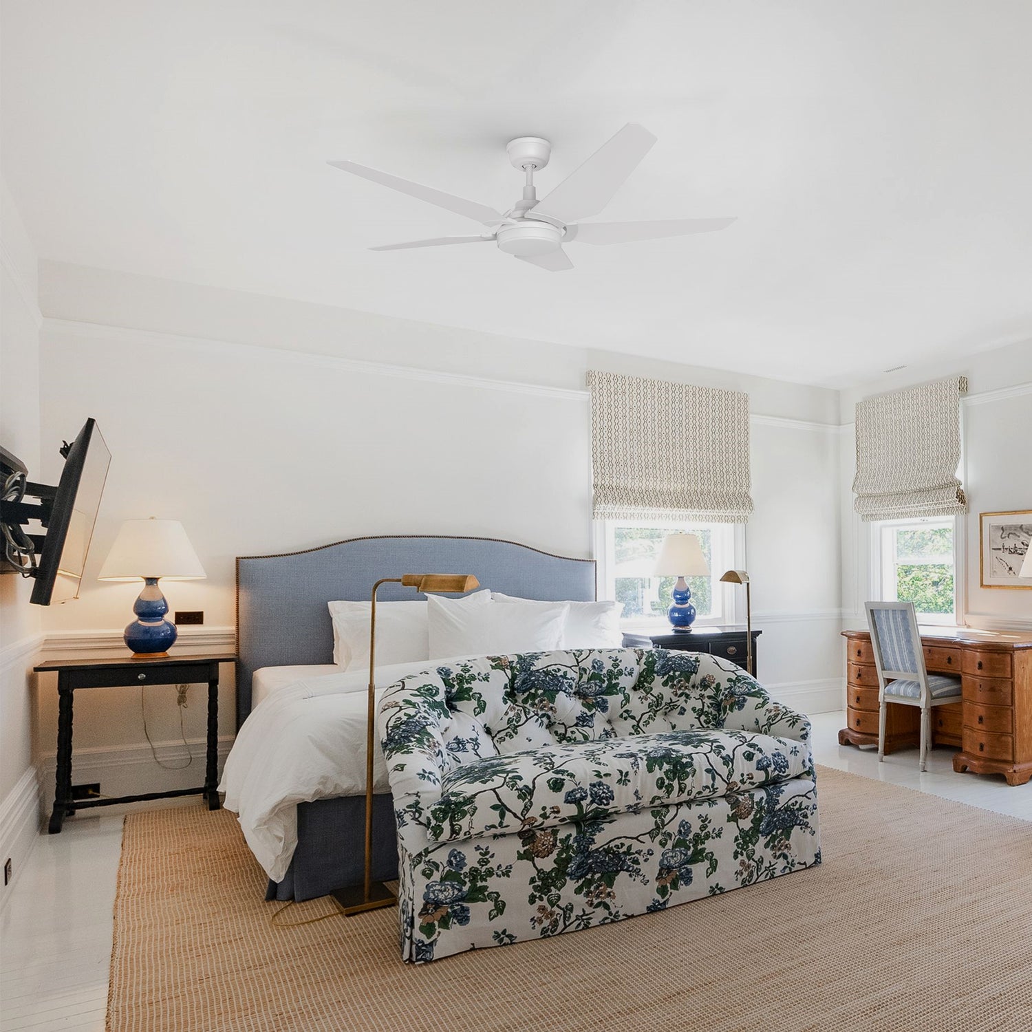 A serene and stylish bedroom features a white Smafan Voyager 52” Smart WiFi ceiling fan in white with light centered on the ceiling. The room is decorated in soft neutrals with a blue upholstered bed, a floral loveseat at the foot, woven blinds, and classic wooden furnishings. Natural light streams in through two large windows, enhancing the calm, cozy atmosphere.