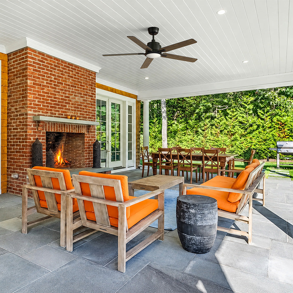 A covered patio with Smafan 52-inch black five-blade ceiling fan, brick fireplace, orange-cushioned seating, and outdoor dining area surrounded by lush greenery.