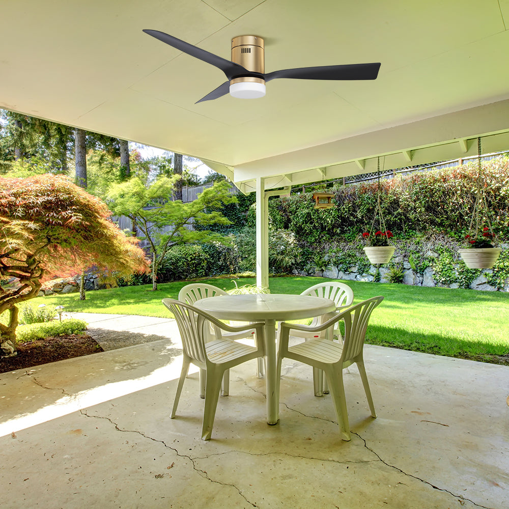 Outdoor patio featuring a 48-inch black and gold low-profile ceiling fan with an integrated light. The fan has three black blades and a gold motor housing, installed on a white ceiling. The patio includes a white round table with four plastic chairs, surrounded by greenery, hanging plants, and a landscaped garden, showcasing the ceiling fan’s damp-rated suitability for outdoor spaces. 