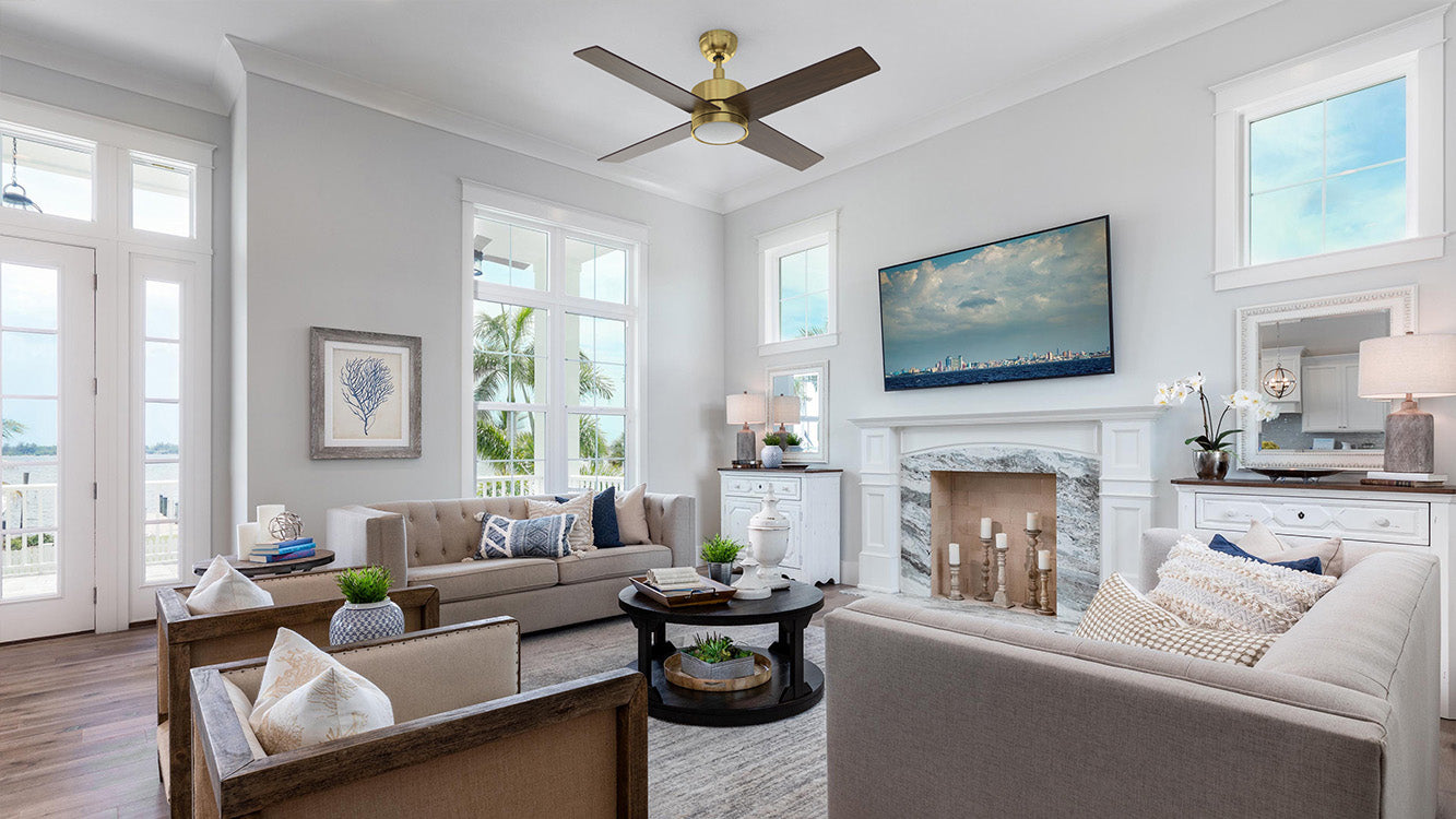 A spacious, sunlit living room is furnished with two beige sofas and two wood-framed armchairs arranged around a round black coffee table. Centered in the room is a 52 inch Smafan Rossendale gold ceiling fan with light, featuring four dark wood blades and a sleek gold motor hub. The room's focal point is a white fireplace with a gray marble surround, topped by a large flat-screen TV. Large windows and a glass-paneled door offer a view of palm trees and water, enhancing the coastal aesthetic.