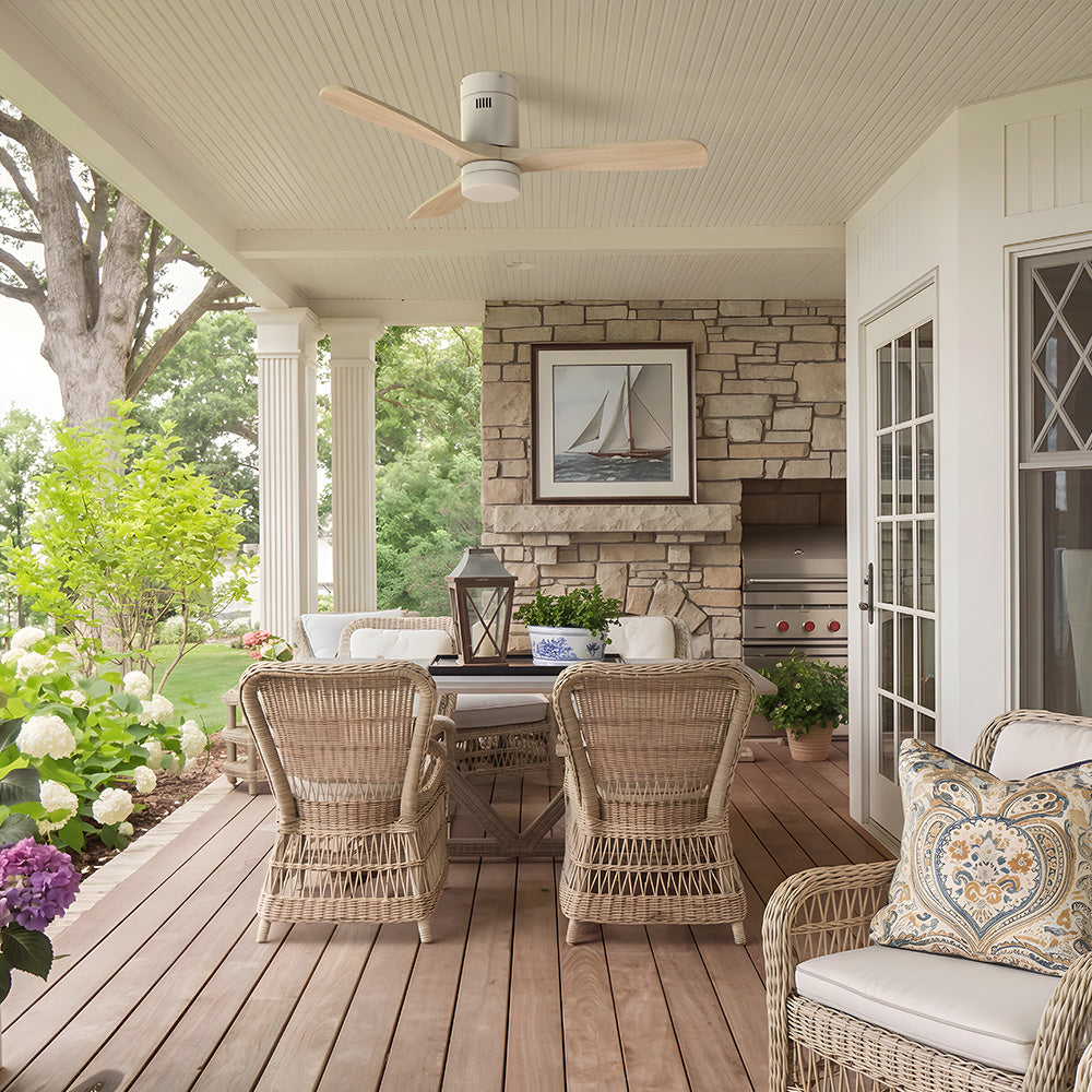 Smafan Alfa 52 inch low profile ceiling fan in white with light wood blades installed on a covered outdoor porch, complementing the coastal farmhouse design with wicker seating, a stone fireplace, and a spacious outdoor dining area. 