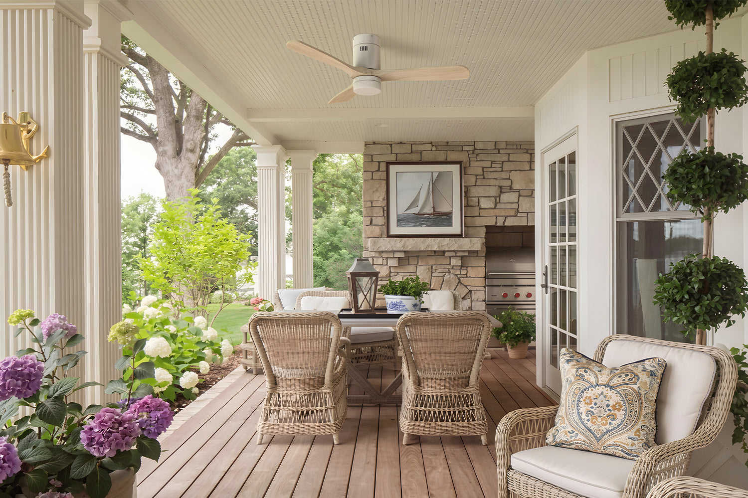 Smafan Alfa 52 inch flush mount outdoot ceiling fan in white with light wood blades installed on a covered porch, complementing the coastal farmhouse design with wicker seating, a stone fireplace, and a spacious outdoor dining area.