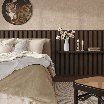 Modern cozy bedroom with a beige and taupe bedding set, featuring a textured wood slat accent wall in dark walnut finish. A floating shelf above the bed holds a minimalist white vase with pink blossoms, two lit candles in sculptural holders, and a small book. The room also includes a round wall art piece with abstract koi fish design, a woven rug, and a cane-seated chair in the foreground, creating a warm and serene ambiance.