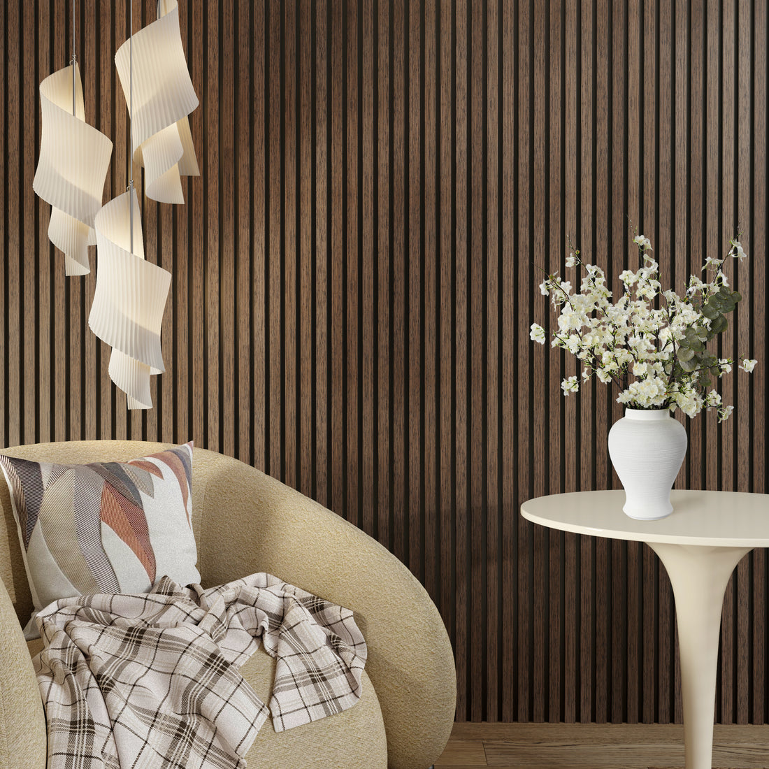 A cozy reading corner featuring an American Smoked Walnut acoustic slat wood wall panel as a rich backdrop. A sculptural cream lounge chair with a patterned pillow and plaid throw sits next to a round pedestal side table holding a white vase of delicate white flowers. Overhead, modern cascading pendant lights add a soft, artistic glow.