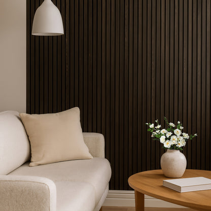 Modern living room featuring an American Smoked Walnut acoustic slat wood wall panel, a light beige sofa with a matching cushion, a white pendant light, and a round wooden coffee table with a ceramic vase of white flowers and a closed book.