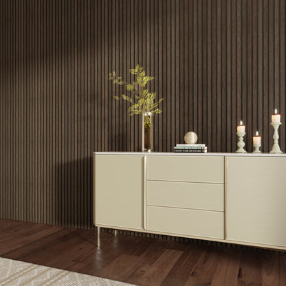 Interior scene featuring an American Smoked Walnut acoustic slat wood wall panel as the backdrop. A sleek beige sideboard stands against the wall, decorated with candles, books, and a tall vase with green branches. The dark wood flooring and vertical slat design add warmth and sophistication to the space.