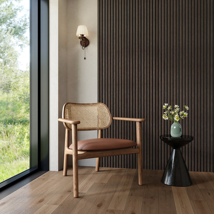 A bright, modern corner featuring a smoked walnut acoustic slat wood wall panel that adds depth and warmth to the space. The setting includes a mid-century rattan lounge chair with a leather cushion, a sculptural black side table topped with a light green vase and fresh daisies, and a small vintage-style wall sconce. Natural light floods in through a floor-to-ceiling window, enhancing the organic textures and earthy tones of the room.