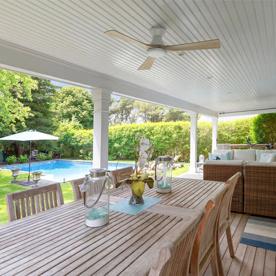 White flush mount wood ceiling fan with light installed on a white covered patio ceiling above a large wooden outdoor dining table with a backyard swimming pool view. #color_light-wood